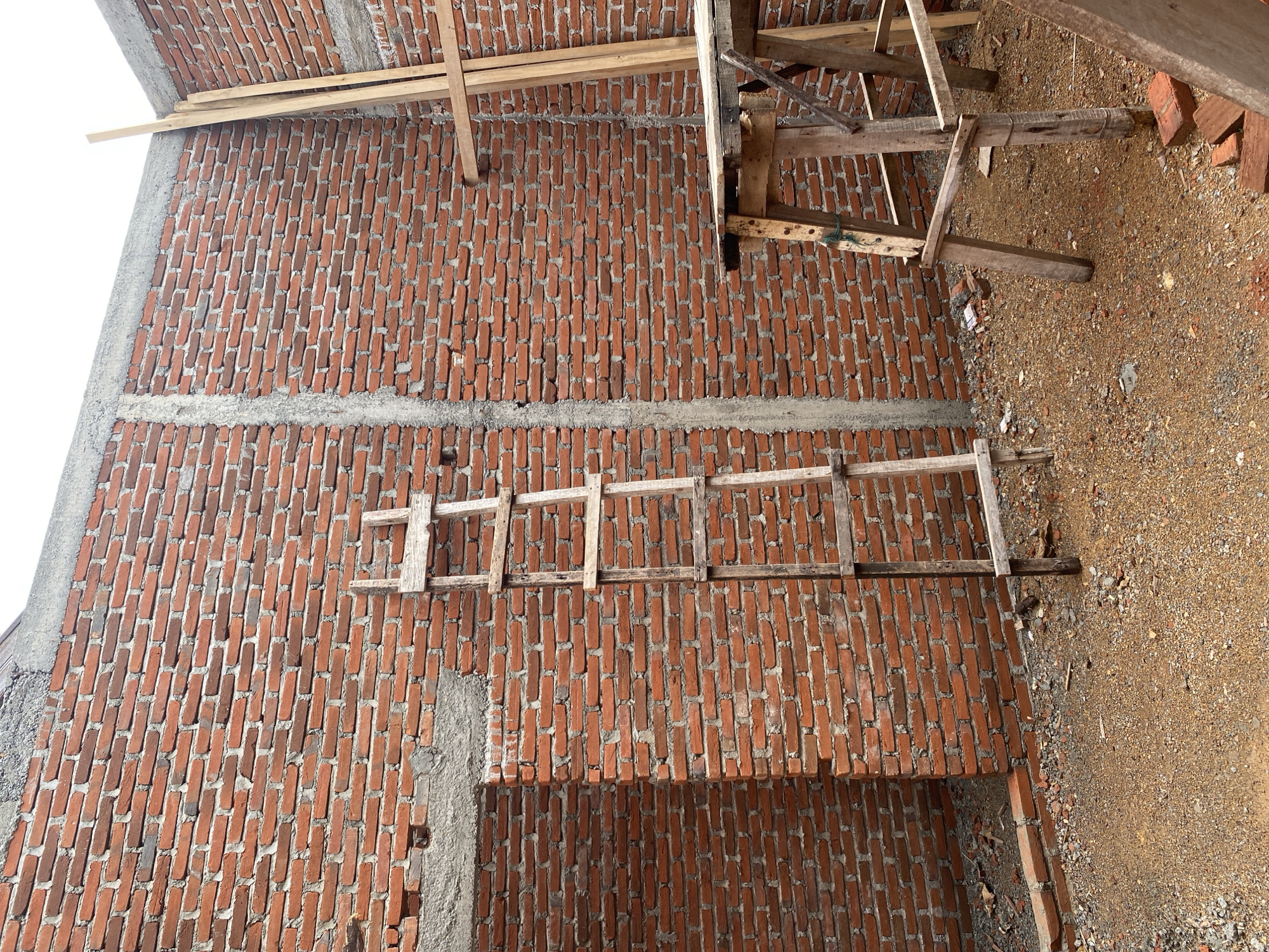 The image shows a construction site with unfinished brick walls. A wooden ladder leans against the wall. In the foreground, there is a wooden scaffolding structure and loose bricks scattered on the gravelly ground. The sky is visible in the background, suggesting it is daytime. The location is identified as Unknown, Unknown.
