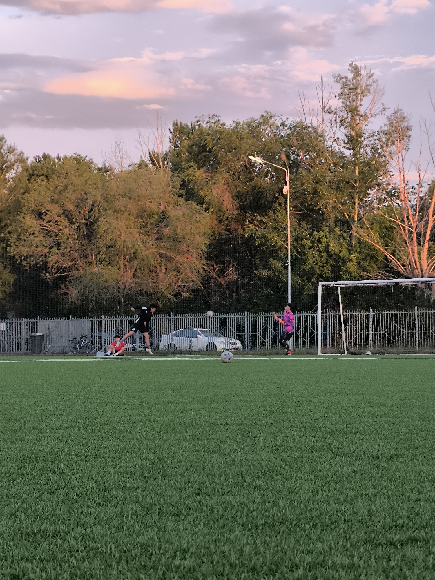 An outdoor scene depicts a soccer field at dusk with a pink and purple sky. In the middle ground, three individuals are visible on the green turf. One person, wearing a black shirt and shorts, is in mid-air, appearing to kick a soccer ball that is suspended in the air. Another person, in a red shirt and shorts, is seated on the ground to the left of the kicking player. To the right, a person in a purple shirt and black pants, likely a goalkeeper, stands near a soccer goal. A white soccer ball is on the ground to the left of the goal. A chain-link fence runs horizontally across the middle ground, separating the field from a line of trees and a street behind it, where a white car is parked. A street lamp is positioned near the trees. Bicycles are also visible leaning against the fence to the left. The overall environment suggests a recreational activity taking place in an urban setting.