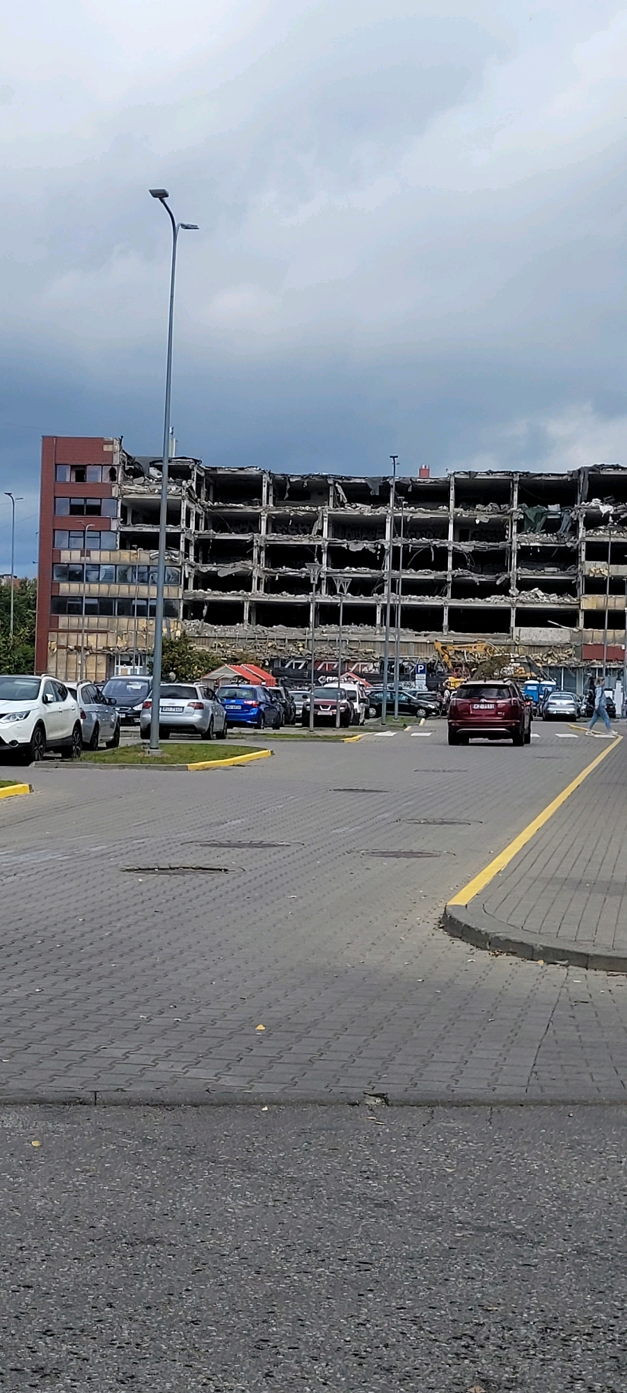 A multi-story building, in a state of partial demolition, dominates the background of this image. The building's facade is heavily damaged, with open floors and exposed structural elements, suggesting ongoing demolition work. A yellow excavator is visible near the base of the building. In the foreground, a paved area, likely a parking lot or street, is lined with parked cars. A red SUV is in motion, driving away from the camera. Pedestrians are also present, walking on the paved area. The sky is overcast with clouds. The scene appears to be set in a commercial or industrial area of Riga, Latvia.