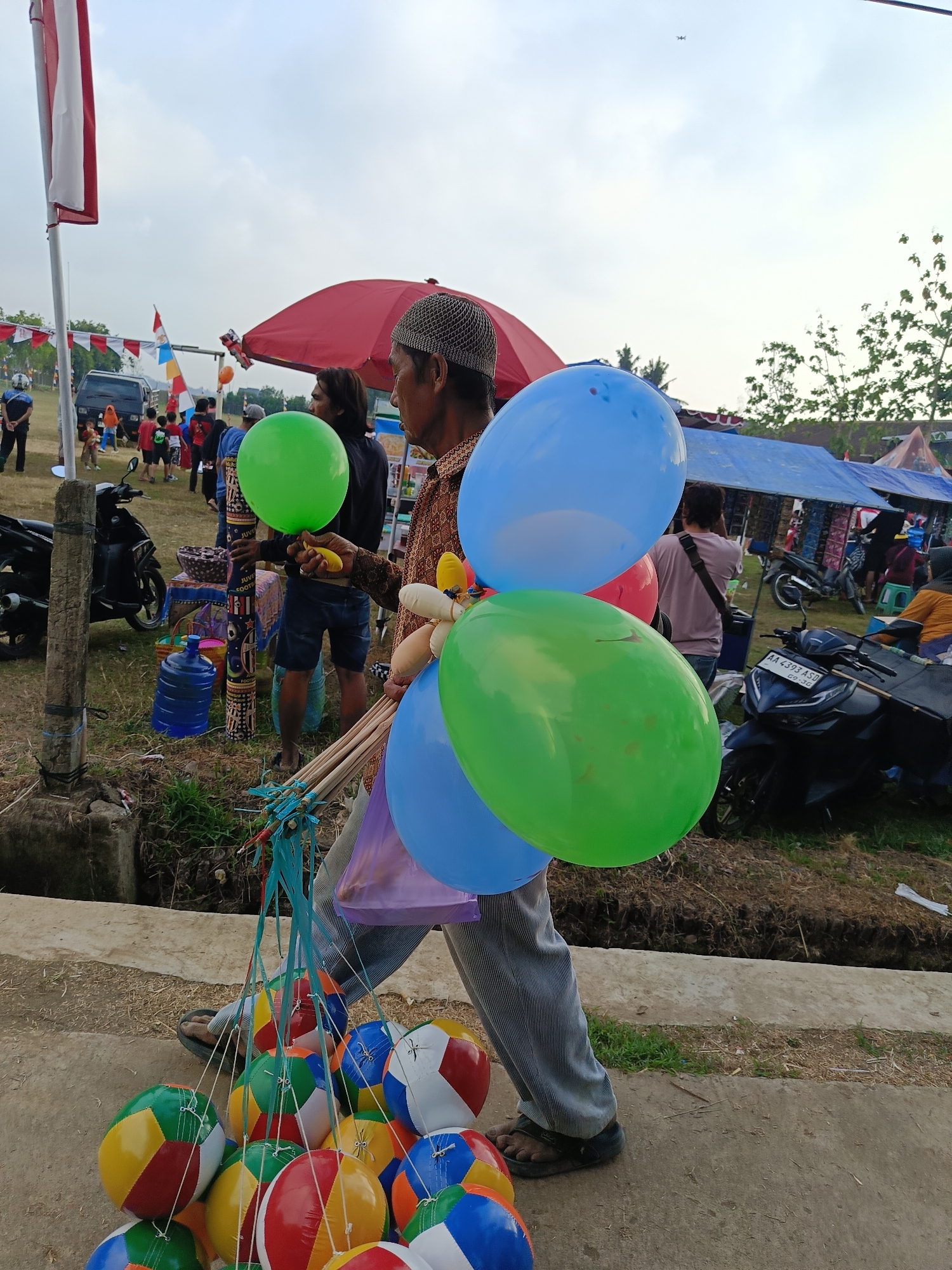 In Mangli, Indonesia, a vendor walks along a dirt path carrying an assortment of balloons and inflatable balls. The vendor, wearing a patterned shirt, a cap, and striped pants, is positioned in the foreground, facing right. He holds a bunch of colorful balloons, including blue, green, and red, by their strings, and has a large cluster of multi-colored inflatable balls dangling from sticks. In the background, a busy market scene unfolds with stalls and people. Several motorcycles are parked, and a red umbrella is set up near some stalls. The sky is overcast, and trees are visible in the distance. A prominent Indonesian flag is displayed on a pole to the left of the image.