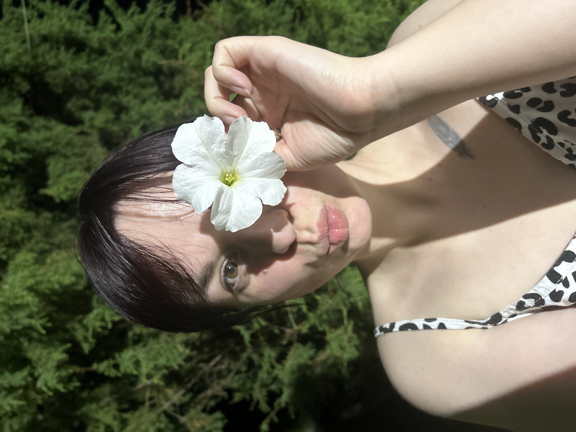 A woman with short, dark hair is holding a white flower in front of her right eye. She is wearing a black and white leopard-print dress. The background is a dark green, out-of-focus hedge. The lighting suggests it is nighttime. The image was taken in the city of Фоки, Russia.
