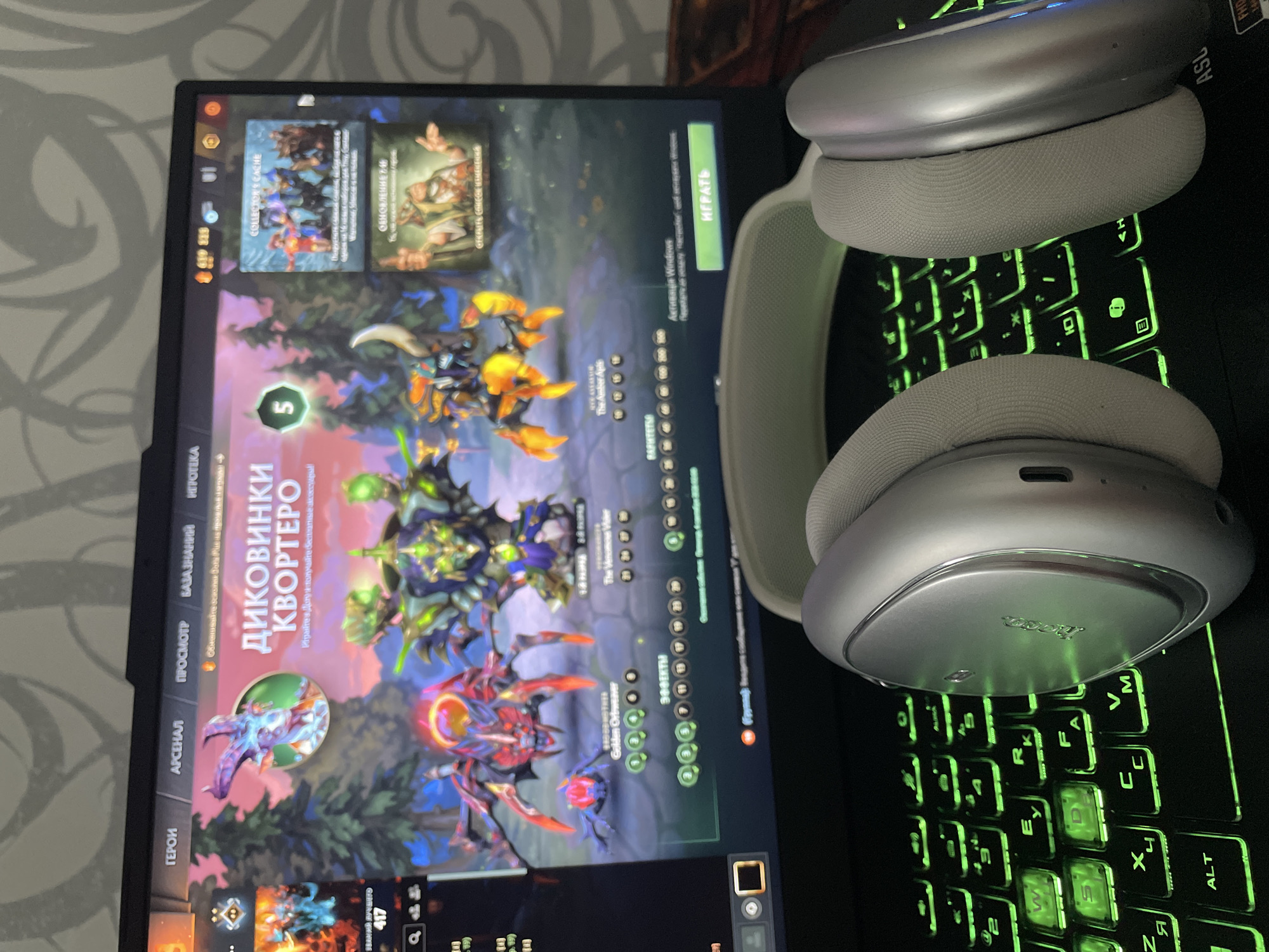 The image shows a close-up of a laptop screen displaying the game Dota 2. The game interface is visible, with character models and text in Russian. To the right of the screen, there is a section labeled "COLLECTOR'S CACHE" and "ОБНОВЛЕНИЕ 7.36". Below the laptop, a pair of silver-colored headphones with light gray earcups rests on a keyboard that is illuminated with green backlighting. The overall scene suggests a person is about to play or is currently playing a video game. The location of Rozsoshentsi, Ukraine is not depicted or suggested in the image.