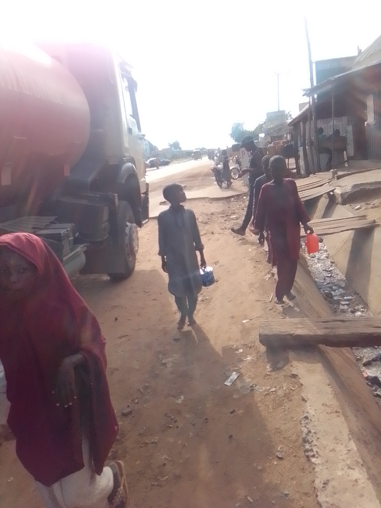 The image captures a dusty street scene in Galadima, Nigeria, with a large tanker truck dominating the left side. The truck's orange tank is partially obscured by bright sunlight. To the right of the truck, a young boy in a dark tunic walks away from the camera, holding a blue bucket. Further down the street, several individuals are visible, including a person in a maroon robe carrying a red container and another person in a dark outfit. In the background, there are wooden structures that appear to be market stalls or shops, and a few motorcycles are parked nearby. The ground is unpaved and covered in dirt and debris. The overall impression is one of a bustling, everyday scene in a Nigerian town.