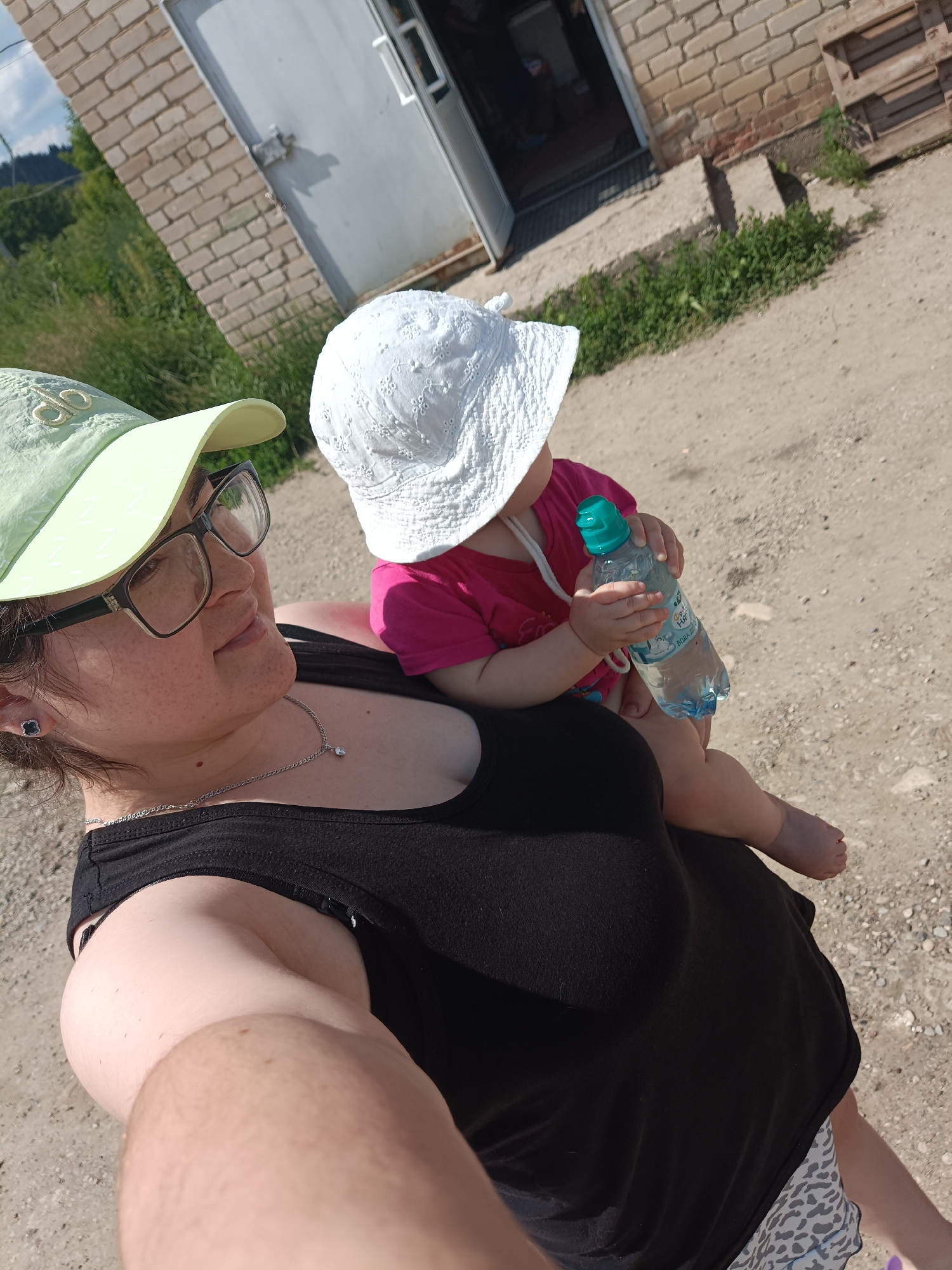 In a close-up, outdoor, selfie-style photograph, a woman wearing glasses, a black tank top, and a light green baseball cap holds a baby in her arms. The baby is wearing a pink shirt and a white sun hat and is holding a bottle of water. The background shows a brick building with a metal door and some greenery. The ground appears to be dirt and gravel. The woman is looking slightly to the right, and the baby is looking away from the camera. The lighting suggests it is daytime, likely sunny. The image appears to have been taken in a rural or semi-rural setting, possibly a backyard or a property near a building. The presence of the water bottle and the attire of the individuals suggest warm weather. There are no clear indicators of the specific location within Posad, Russia, but the overall scene depicts a casual, everyday moment.