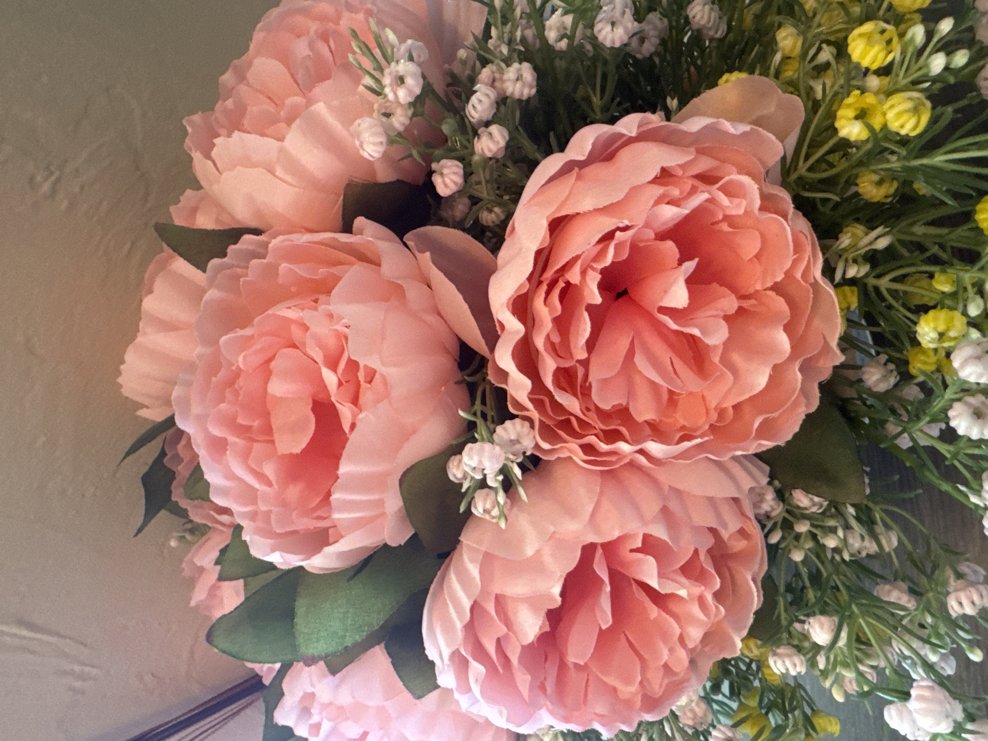 A close-up, top-down view shows a floral arrangement of pink peonies and small white flowers, interspersed with green foliage and tiny yellow blossoms. The peonies are large and lush, with multiple layers of ruffled petals. The arrangement is likely indoors, as indicated by the soft, diffused lighting and the presence of a textured wall or surface in the background, which appears to be a neutral, light beige color. No specific location in Horishni Plavni, Ukraine is discernible from the image.