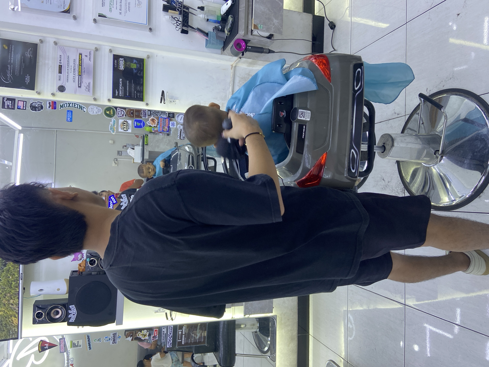 In Kecamatan Pati, Indonesia, a barber with dark hair, wearing a black t-shirt and black shorts, is giving a haircut to a young child seated in a car-shaped barber chair. The child is wearing a blue cape. The barber is holding a black electric trimmer and is actively cutting the child's hair. The environment appears to be a barbershop, with shelves holding certificates, speakers, and various barber tools like scissors and a hairdryer visible in the background. The floor is tiled with a reflective surface.