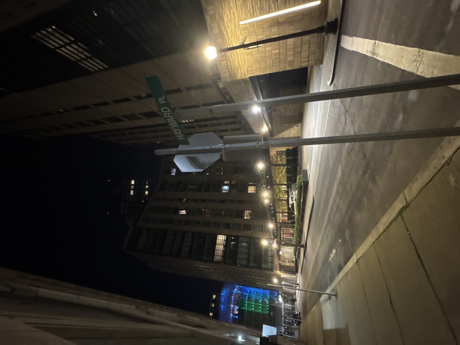 The image displays a nighttime urban street scene on Hayward Place in Boston, Massachusetts, United States. The perspective is low-angle, looking down the street framed by tall, multi-story buildings.
In the immediate foreground, two metallic posts support a green street sign with "HAYWARD PL" in white text, angled towards the right. A blank, silver-gray rectangular sign is affixed to the left post. On the right, a black ornate lamppost with a warm-toned globe light illuminates the concrete sidewalk and a building with a textured, light-colored facade featuring vertical strip lighting.
The asphalt street appears dark and exhibits wet patches, reflecting ambient light. Faded white painted lines are visible on the street surface. On the left, a concrete sidewalk runs parallel to the street. Further down the left side, a multi-story building with illuminated windows is present, beyond which another building facade glows with distinct blue and green light. Two light-colored sedans are parked parallel to the curb under an awning or covered entrance on the left, illuminated by a series of smaller, warm-toned lights. No individuals are present in the scene. The sky is dark and clear.