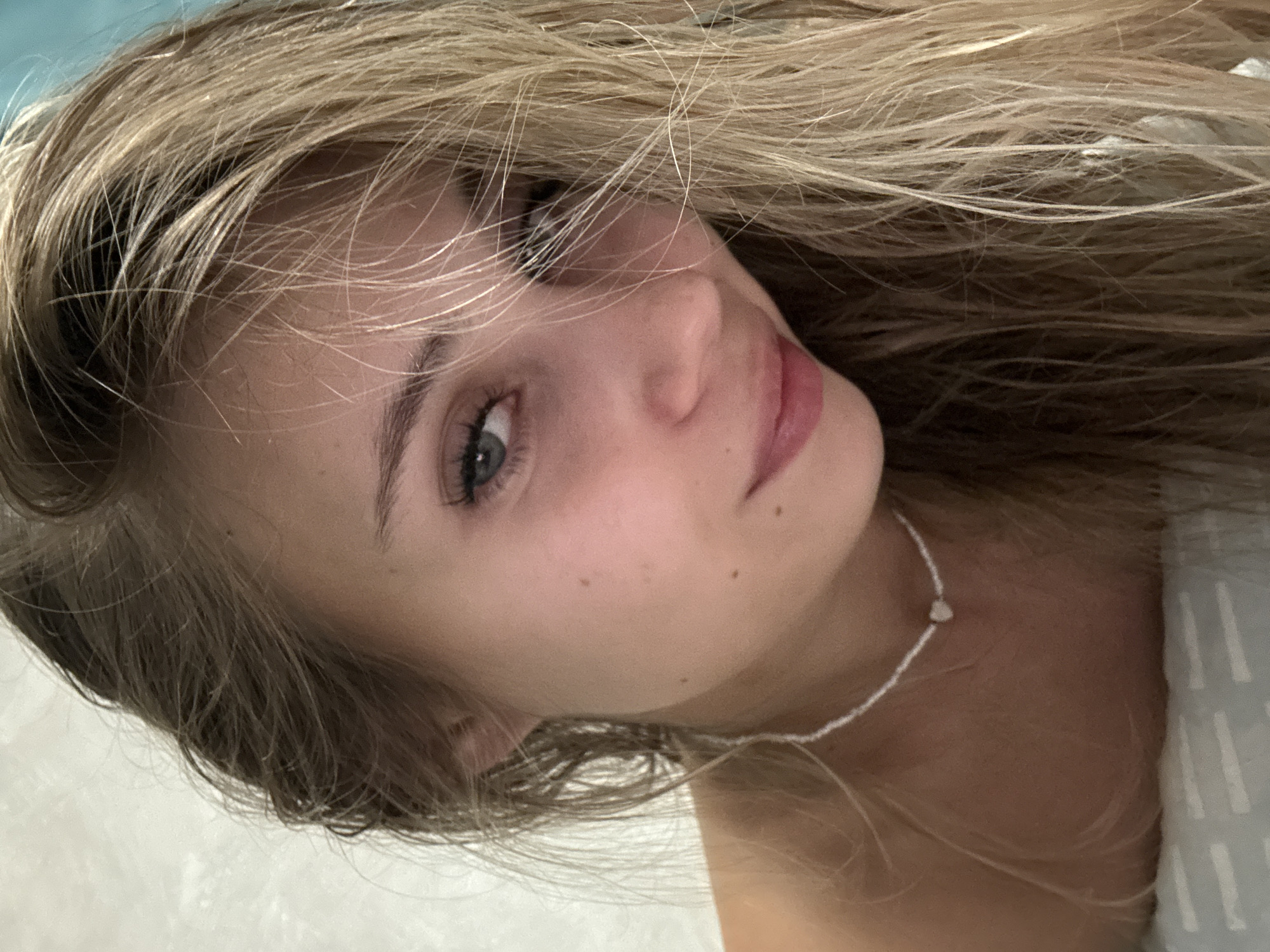 Close-up portrait of a young woman with light blonde hair partially obscuring her face. She has fair skin, gray eyes, and full lips. She wears a white beaded necklace. The background is indistinct but appears to be a light-colored surface, possibly a bed sheet. The image is tilted. There is no information in the image to determine if this photo was taken in Chaikovsky, Russia or elsewhere.
