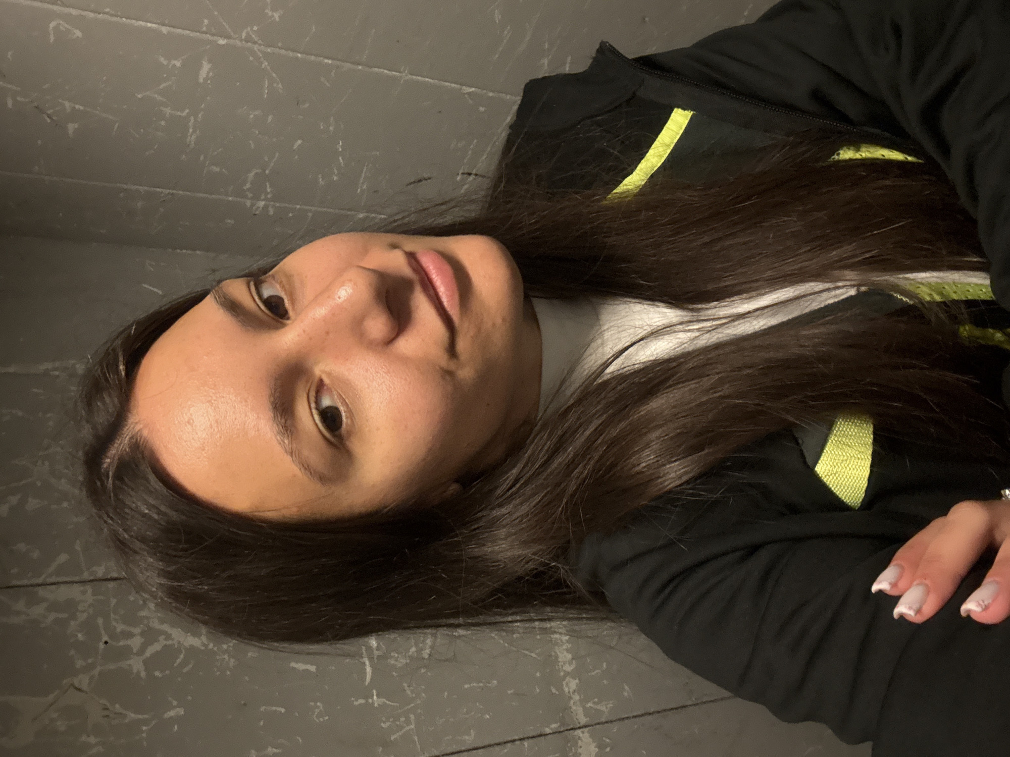 A young woman with long, dark brown hair is pictured indoors against a gray-toned textured wall, reminiscent of concrete. She's wearing a black jacket with light green accents over a white turtleneck sweater. Her face is partially visible, showing smooth skin and dark eyes. Her right hand, with manicured nails, rests on her jacket. The image is taken at an angle, not perfectly straight on. There's no way to know the location in the city of Varna, Bulgaria from the image alone.