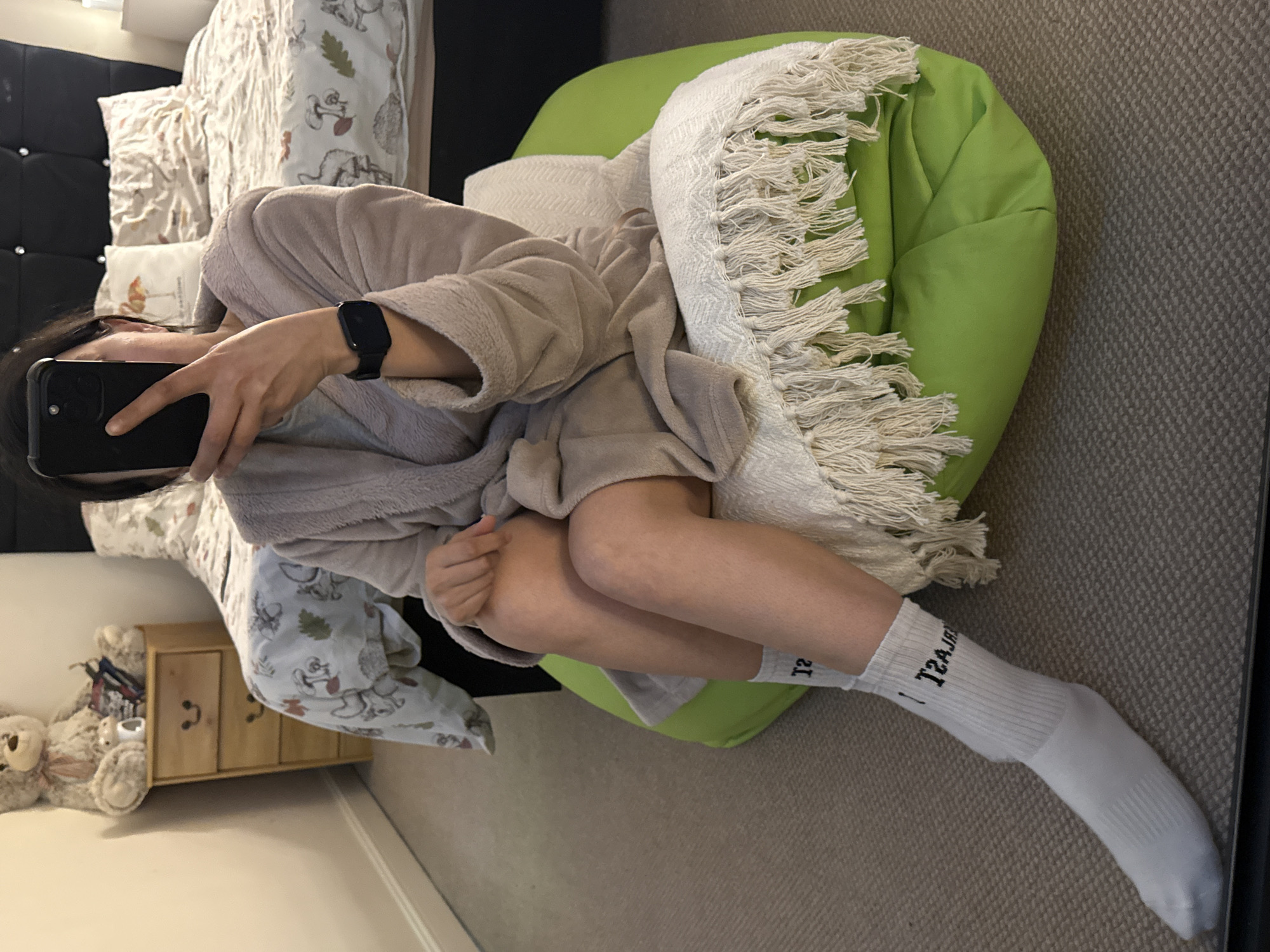 A person wearing a robe and white socks is seated in a green beanbag chair, taking a selfie with a black smartphone. The person is positioned in a room with a bed featuring a black headboard and patterned bedding, a wooden bedside table with a teddy bear next to it, and gray carpeting. The socks have text written on them. A white blanket with tassels is draped on the beanbag. The location is a room in a residence, likely in Great Yarmouth, United Kingdom.
