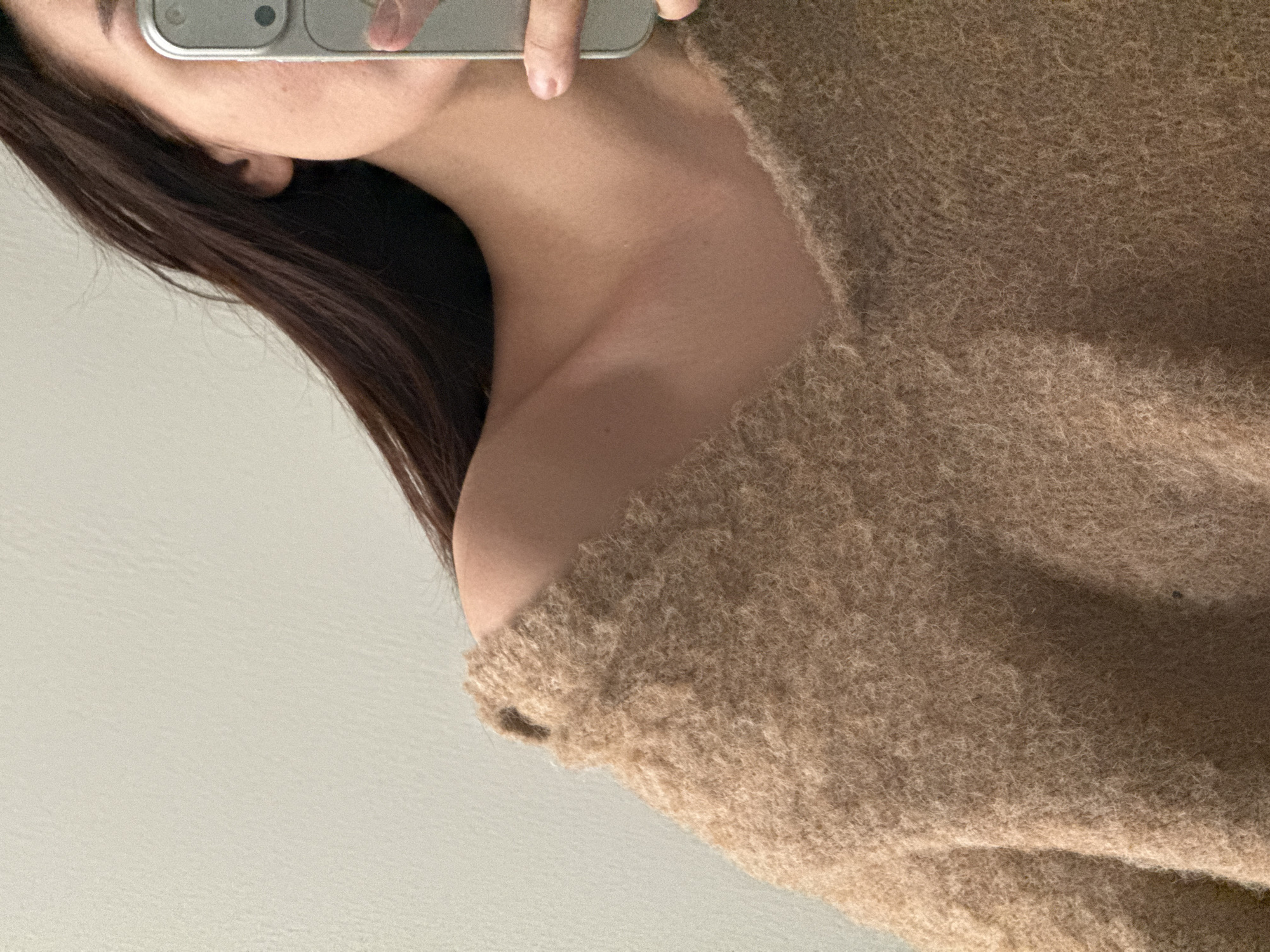 A selfie captures a woman's left shoulder and neck, draped with a beige, fuzzy sweater. The woman's face is partially obscured by a silver cell phone she holds to take the picture. Her brown hair falls along the side of her face and shoulder. The background appears to be a plain, light-colored wall. There is no indication of the location being Chaikovsky, Russia, or any other specific city.