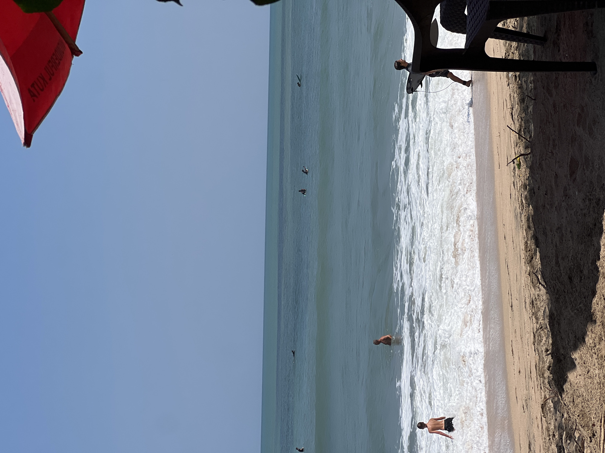 The image depicts a beach scene in Kuta, Indonesia. The ocean, a vibrant turquoise and blue, stretches to the horizon under a clear, pale blue sky. Gentle waves break on the sandy shore, creating frothy white foam. Several surfers are visible in the water, some riding waves and others paddling on their boards. On the beach, a young person in dark shorts stands in the shallow water, facing away from the camera. Further out in the water, an adult male with light-colored shorts stands facing the shore. In the foreground, a dark plastic chair is partially visible, casting a shadow on the sand, suggesting a shaded area or a cafe. A bright red umbrella is seen at the top right corner of the frame, adding a pop of color to the scene. The overall atmosphere is one of leisure and activity on a sunny day at the beach.