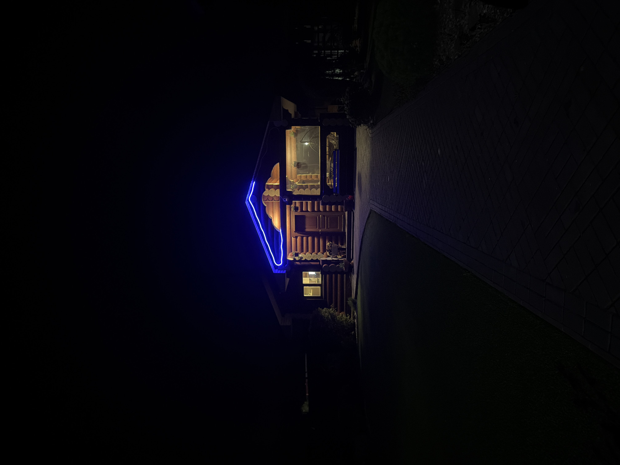 At night, a view of a wooden house in Chaikovsky, Russia. The house features a neon blue outline of its roof, and some lights are visible inside. A brick path leads towards the house, with grass on either side. The surroundings are mostly dark.
