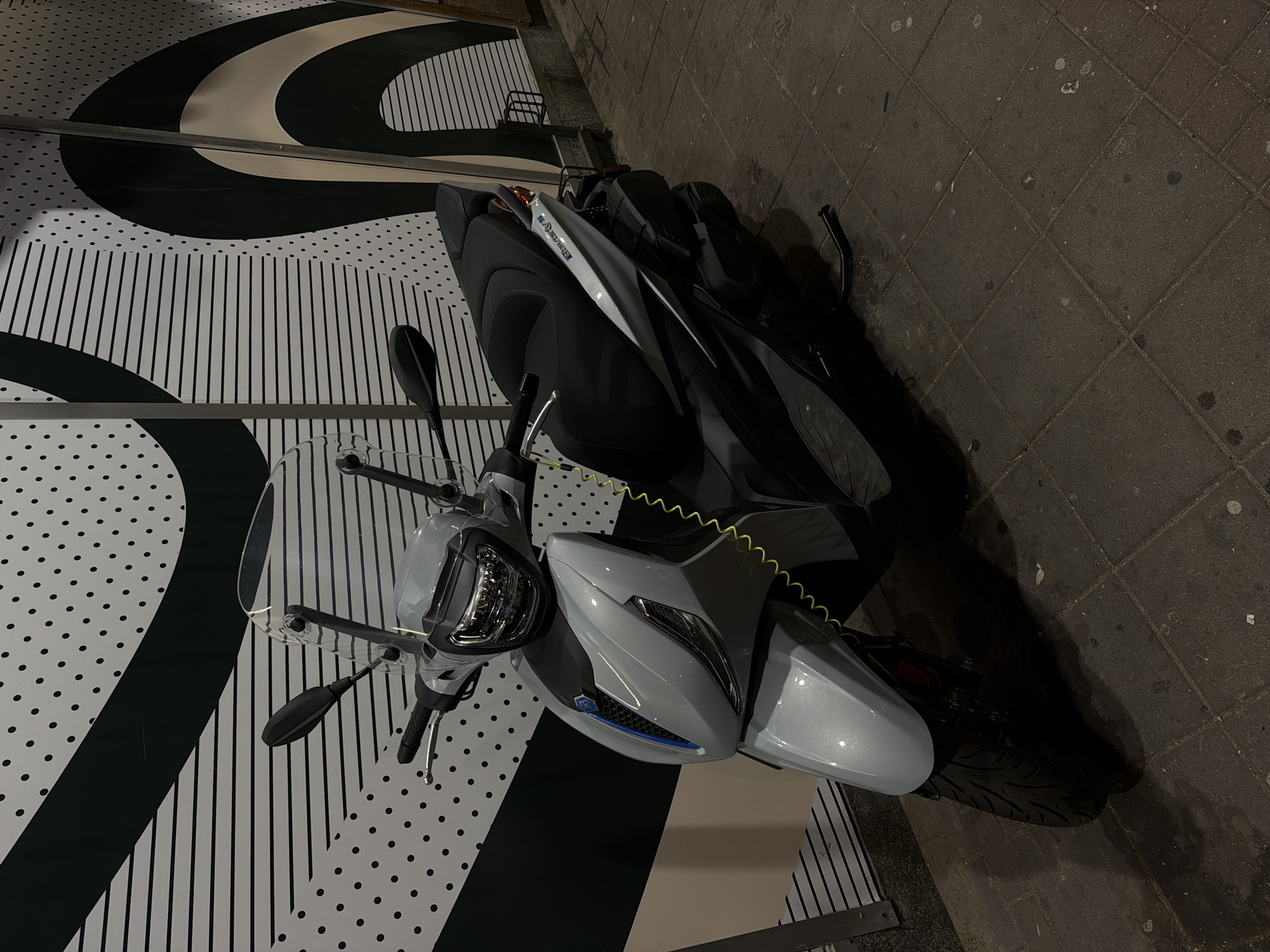 A light gray scooter is parked on a sidewalk in front of a wall with a black and white striped and dotted pattern. The scooter has a black seat, a clear windshield, and a yellow coiled cable attached to its front. The sidewalk is paved with gray square tiles. The scene is illuminated by artificial light, casting shadows on the ground. The location is identified as Vracar Urban Municipality, Serbia. No people are present in the image.
