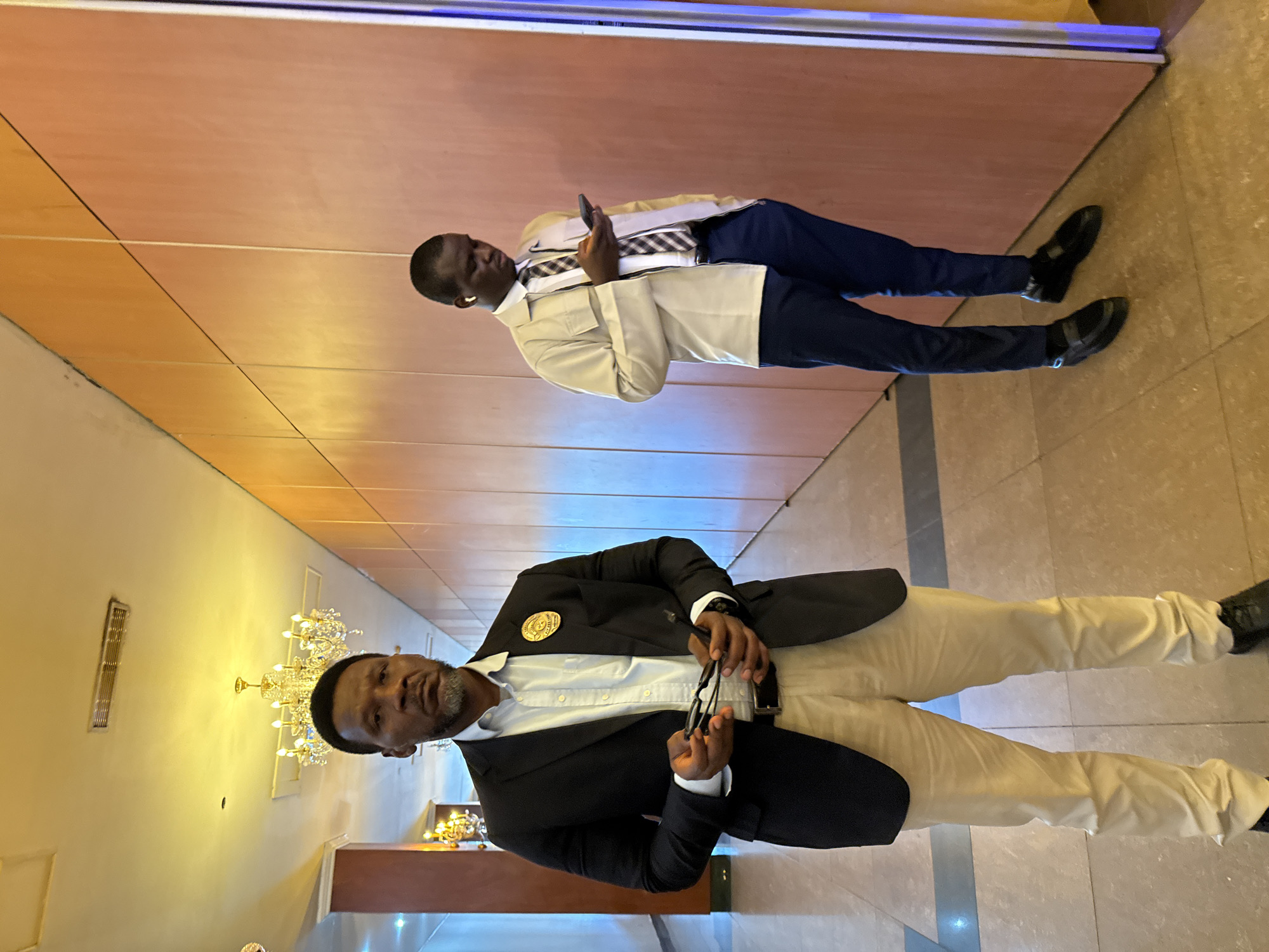 Two men are standing indoors. One is closer to the camera, wearing a black suit jacket, a white shirt, khaki pants, and black shoes. He is holding a pair of glasses in his hands. The other man is standing further away, wearing a tan jacket, a white shirt with a patterned tie, navy blue pants, and black shoes. He is holding a phone in his hand. They are standing in a hallway with light-colored walls and a tiled floor. A chandelier is visible in the background. The location is Itirin, Nigeria.