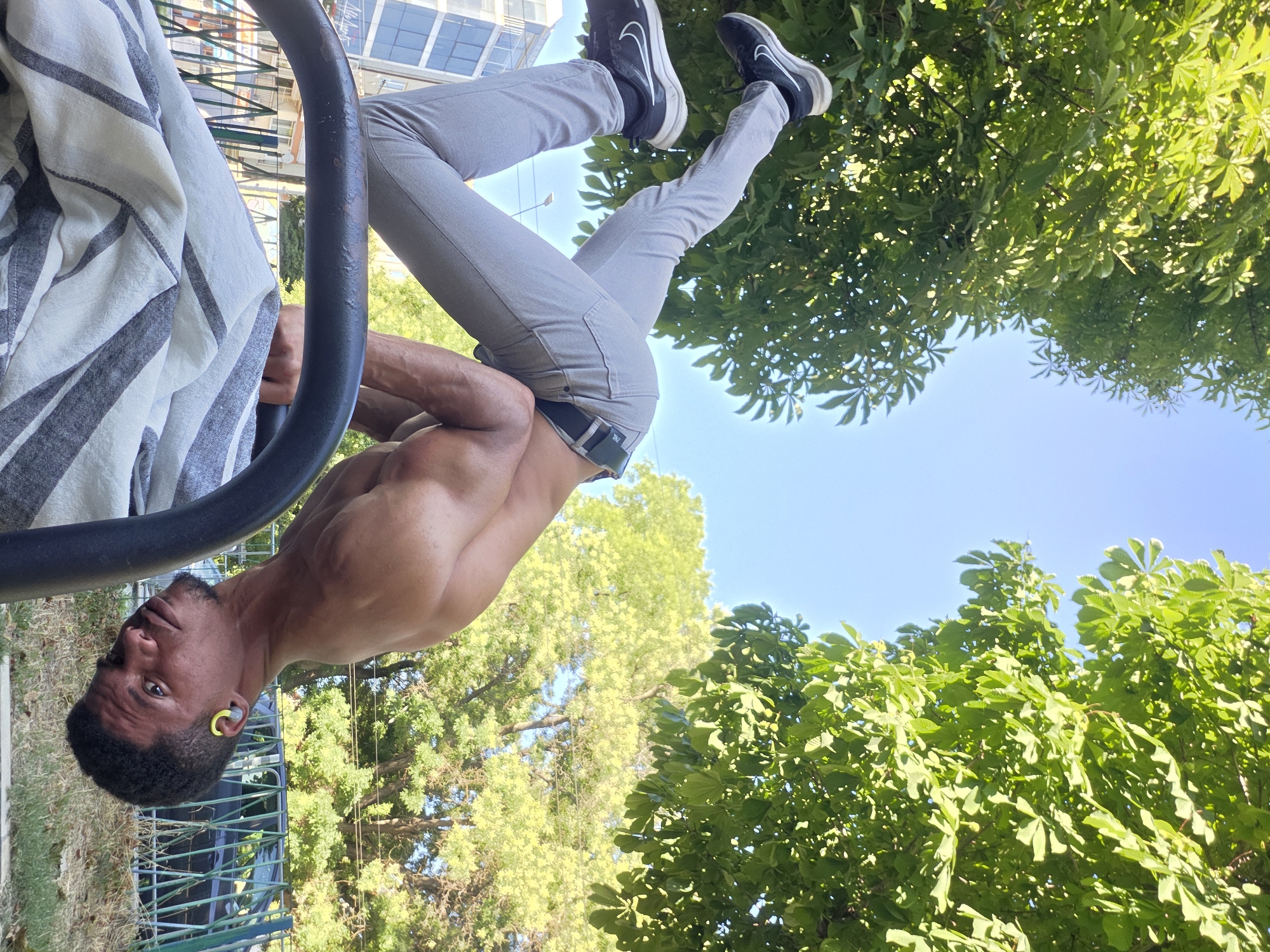 A man is performing a handstand outdoors in Varna, Bulgaria. He is positioned on a horizontal bar, with his body inverted and his feet in the air. The man is wearing light gray pants and black sneakers. His upper body is bare. In the background, lush green trees and a clear blue sky are visible. A building and a fence are also present. The lighting suggests it is daytime, with sunlight casting shadows.