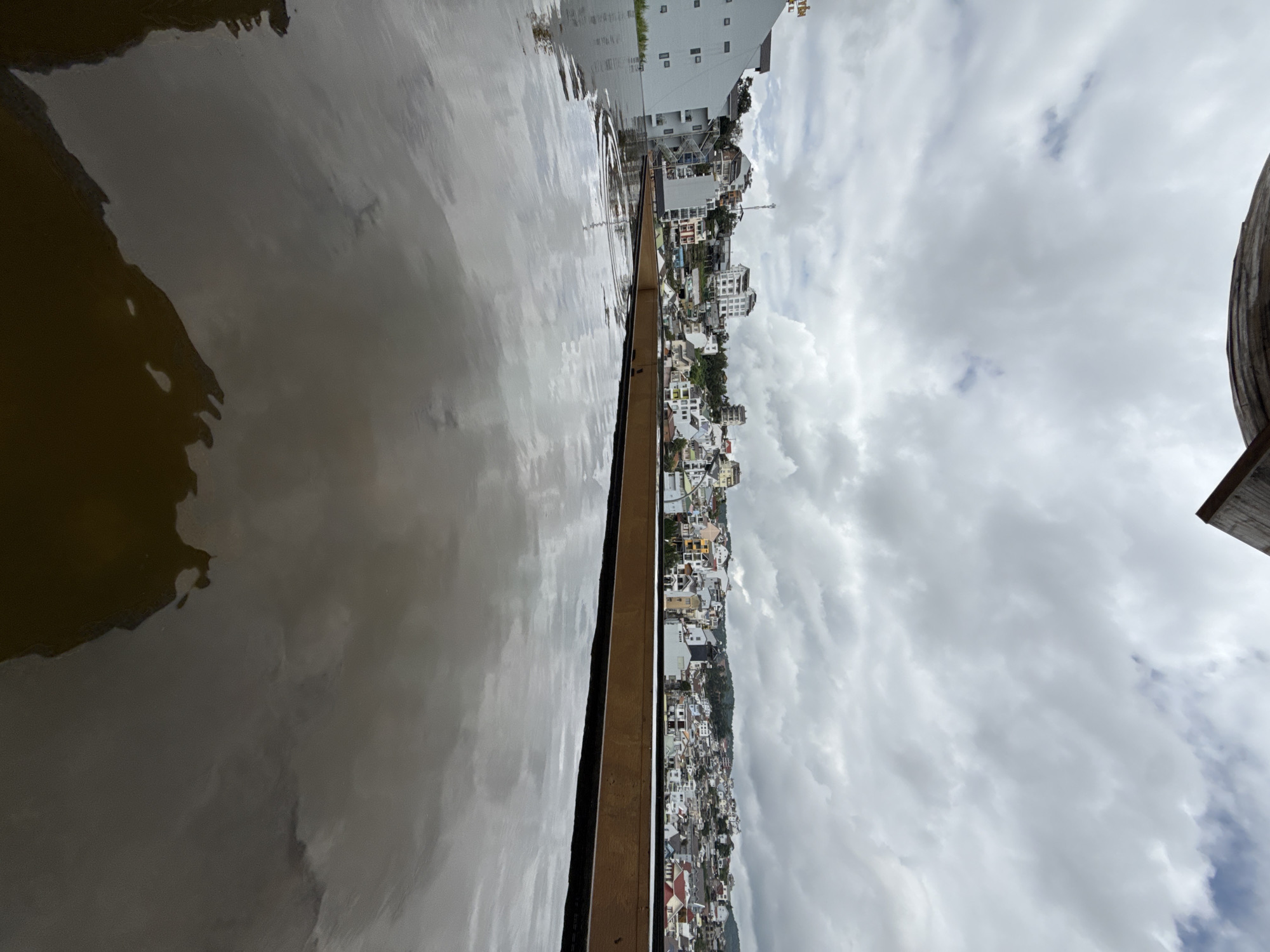 The image shows a cityscape in Da Lat, Vietnam, viewed from an elevated perspective, likely a rooftop. The foreground is dominated by the reflective surface of a pool, possibly an infinity pool, with the ripples on its surface mirroring the cloudy sky and the distant buildings.
The city itself is spread across rolling hills, characterized by a dense collection of buildings with varied architectural styles. Many structures appear to be residential or small hotels, with pitched roofs and a mix of white, pastel, and earthy colored facades. The architecture suggests a blend of traditional Vietnamese and colonial influences. Some taller, more modern buildings are also visible, including a grey multi-story building on the left side of the frame. A telecommunications tower can be seen in the distance, rising above the residential buildings.
The sky is overcast with heavy grey clouds, indicating a potentially rainy or gloomy day. The overall impression is one of a densely populated urban area nestled within a hilly landscape. The water in the foreground creates a striking contrast between the man-made structure and the natural elements of the sky and landscape, with the reflections blurring the lines between them. There are no people visible in the image, and no specific actions or interactions are depicted. The scene is static and focuses on the visual composition of the city and the reflective pool.