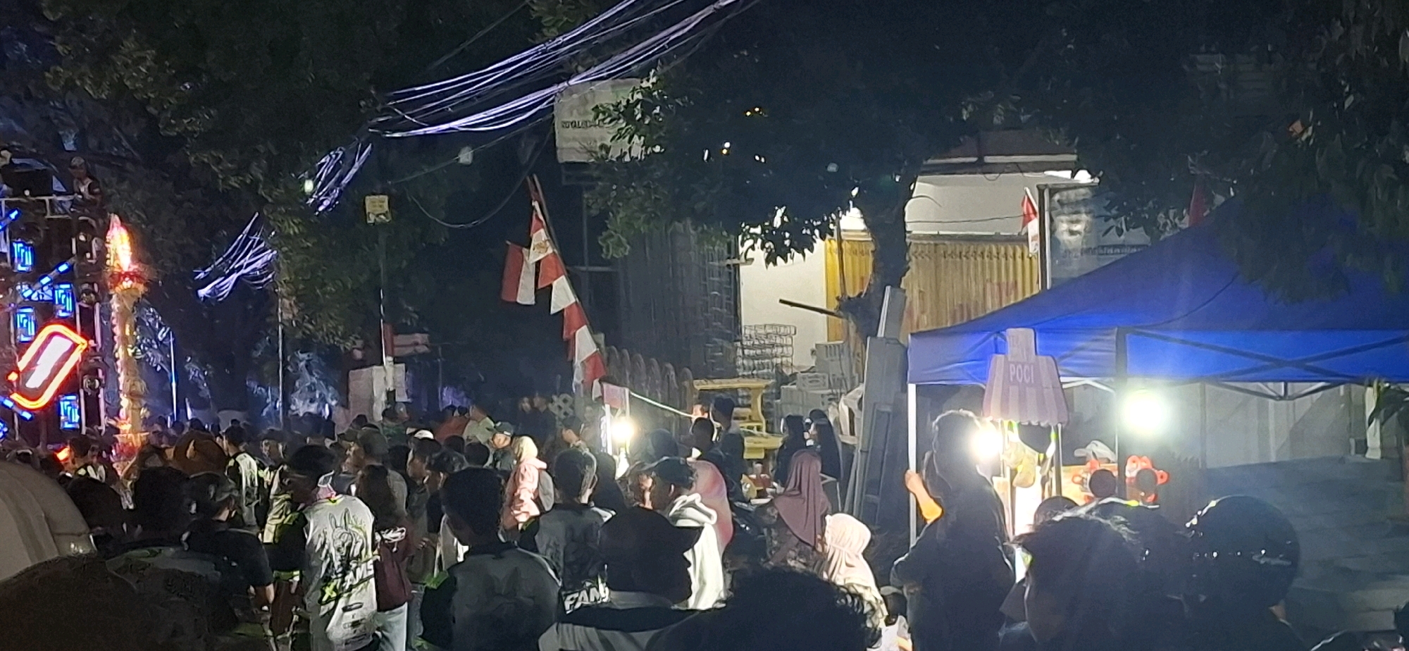 This image captures a lively night scene in Prigen, Indonesia, likely during a local event or gathering. A crowd of people is visible, many of them young, dressed in casual attire. Some are wearing t-shirts with graphic designs.
Dominating the left side of the frame is a tall, illuminated structure, possibly a stage or an elaborate sound system, adorned with bright blue and red lights. To the right, a blue canopy tent is set up, serving as a stall or booth. A sign with the word "POCI" is attached to a decorative light fixture above the tent.
Several flags are present. One is a prominent red and white striped flag, possibly a national flag, displayed on a pole. Another dark flag with white stripes appears to be blowing in the wind.
The environment is dark, with the scene illuminated by artificial lights from the structures and possibly streetlights. Trees with dark foliage overhang parts of the scene, contributing to the nighttime atmosphere. The background shows buildings and fences, suggesting an urban or semi-urban setting. The overall impression is one of a festive and bustling community event.