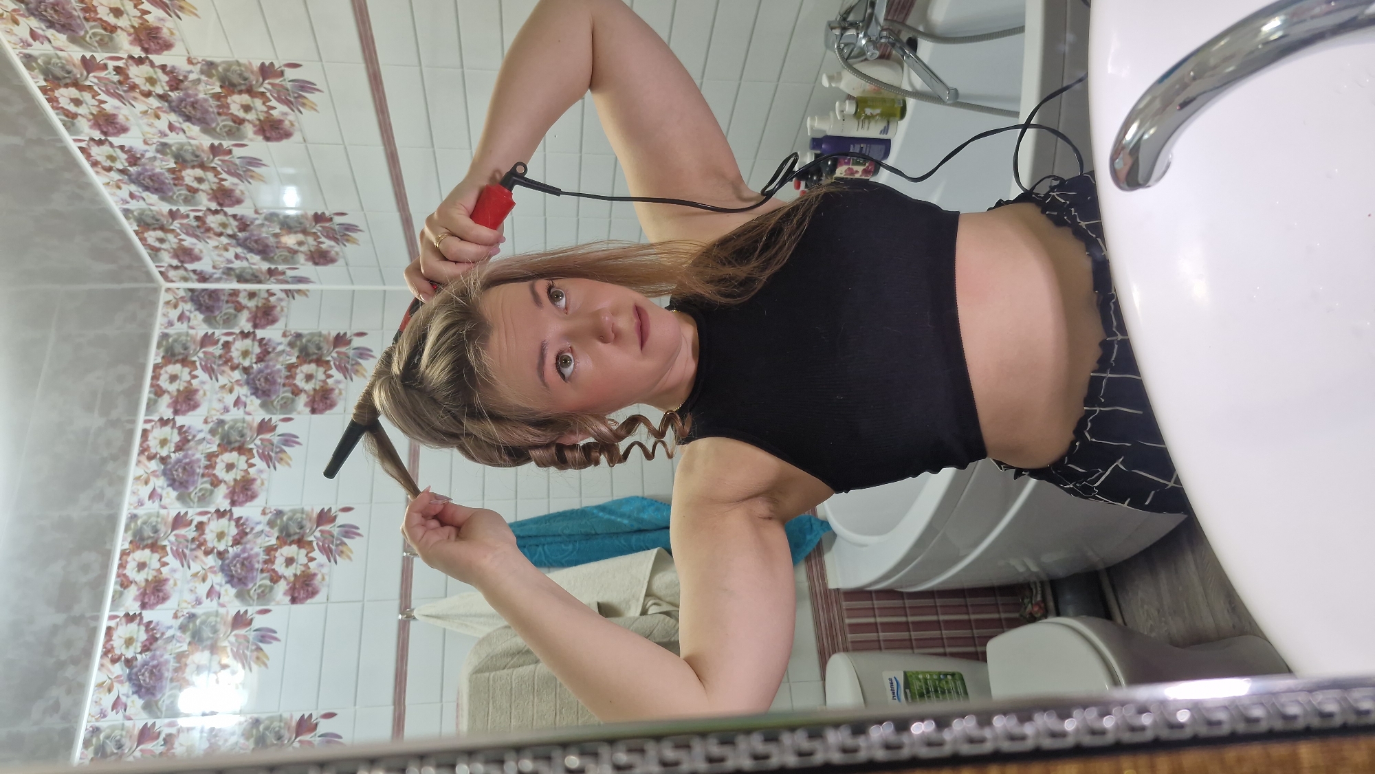 A woman is captured in a mirror selfie, in the process of curling her hair within a bathroom, likely in Otchor, Russia. She has long, light brown or blonde hair, with one side already formed into loose curls. She holds a red and black hair curler/iron in her right hand, coiling a section of hair around it, while her left hand is positioned near the end of the strand. She is wearing a black ribbed sleeveless top and patterned dark pants with a white geometric design. Her gaze is directed towards the camera with a neutral expression.
The bathroom setting includes white tiled walls, with a prominent section featuring a floral pattern in purple, pink, and green hues. A white sink with a chrome faucet is visible on the right, and behind it, various bottles of toiletries are arranged on a shelf. An electrical cord from the hair curler extends towards this area. A white toilet is partially visible below the sink. The floor appears to have light-colored, possibly wood-look, tiles, and a striped mat is also present. A blue/teal towel and a beige towel are visible in the reflection. On the floor, a white bottle with the visible text "naturaS" is seen. The overall scene suggests a routine personal care activity.