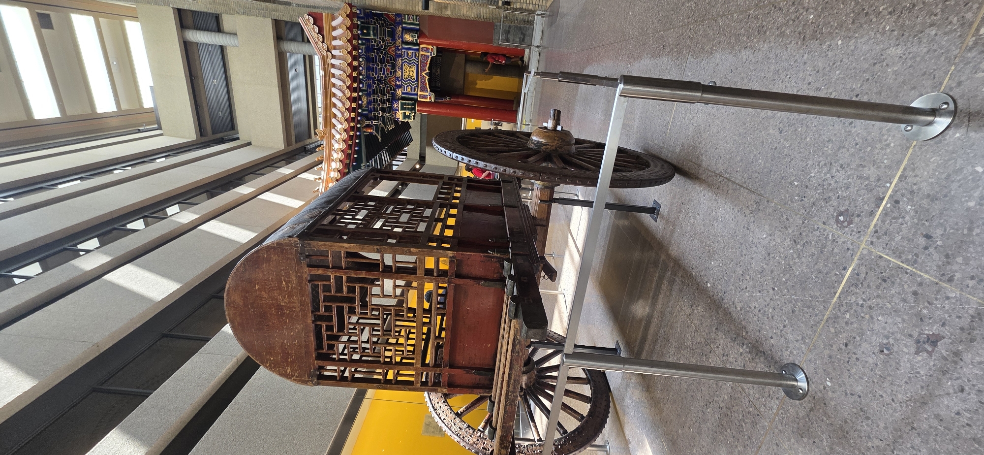 The image shows an ornate, wooden cart with decorative latticework on its sides and a curved wooden roof. It is supported by two large wooden wheels with spoked rims. The cart is positioned in what appears to be a museum or exhibition hall. In the background, there is a colorful, traditional-style structure with intricate carvings and vibrant colors, possibly representing an architectural element from East Asia. The floor is a speckled terrazzo with brass inlays. Sunlight streams in from above, casting shadows and highlighting the textures of the objects. Two metal support poles with circular bases are visible, holding up a horizontal metal bar that runs in front of the cart, suggesting it is part of an exhibit. The overall scene conveys a sense of historical display and cultural exhibition. The lighting suggests it is daytime. There is no visible text in the image.