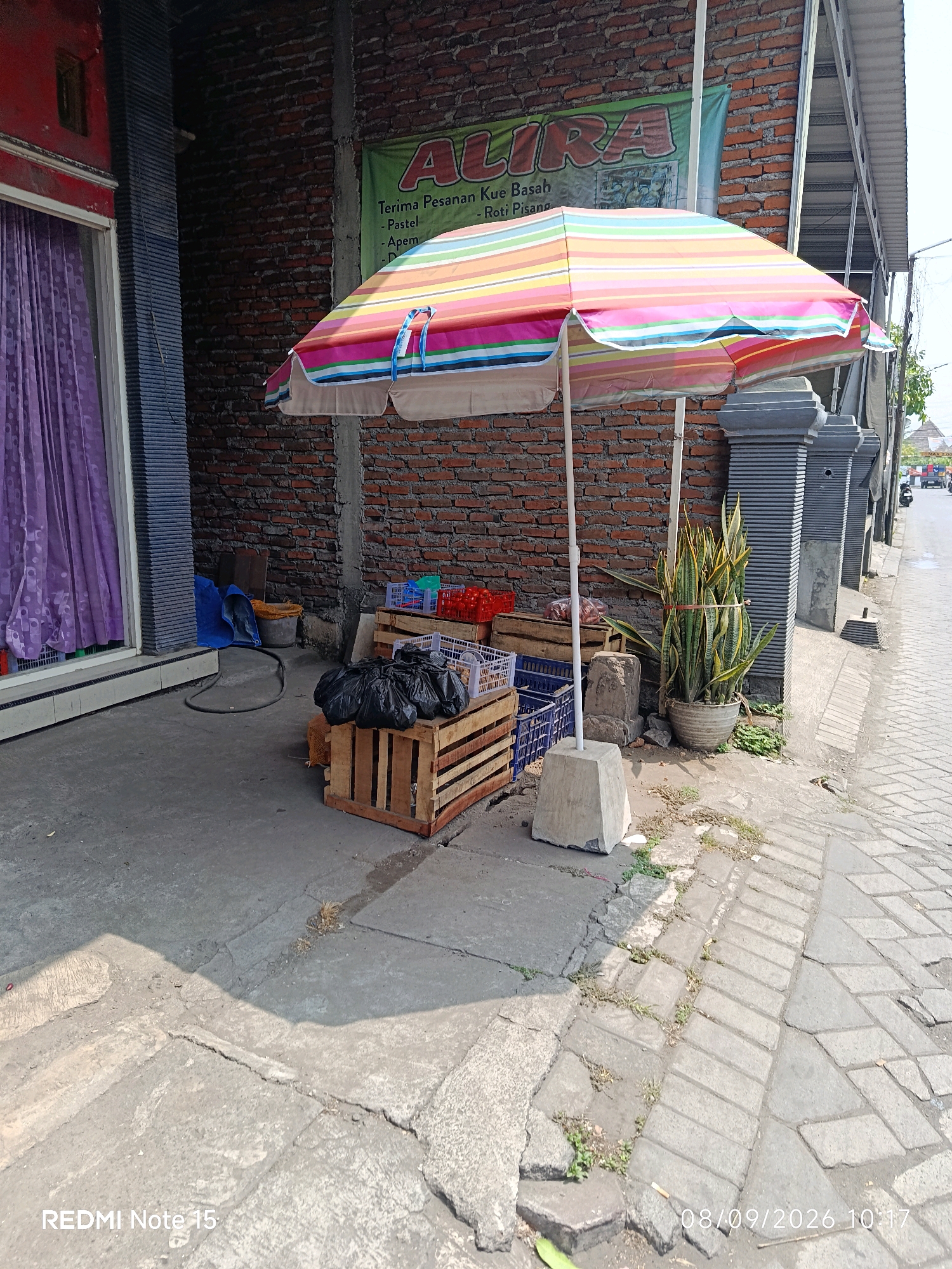 The image shows a street scene in Keboansikep, Indonesia. In the foreground, a colorful umbrella is set up, casting a shadow on the cracked concrete ground. To the left of the umbrella, wooden crates are stacked, with black trash bags and some food items placed on them. Behind the crates, a tall snake plant in a pot stands beside a brick wall. A banner with the text "ALIRA" and some Indonesian writing is visible on the brick wall. To the right of the plant, a sidewalk made of paving stones runs alongside the street, where a few motorbikes can be seen in the distance. The overall environment appears to be an outdoor marketplace or a roadside stall. The timestamp on the image indicates it was taken on 08/09/2026 at 10:17.