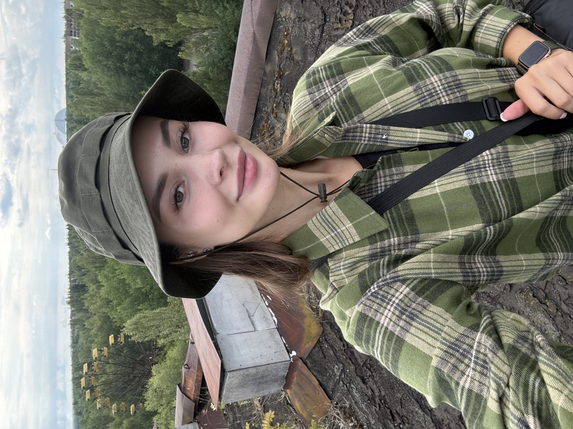 A young woman with brown hair, wearing a green plaid shirt, a bucket hat, and a black watch, stands on a dilapidated rooftop in the city of Pripyat, Ukraine. She is looking at the camera and smiling. In the background, a Ferris wheel, surrounded by trees, and a large, arched structure are visible. The rooftop appears to be made of rusty metal and concrete. The sky is overcast.