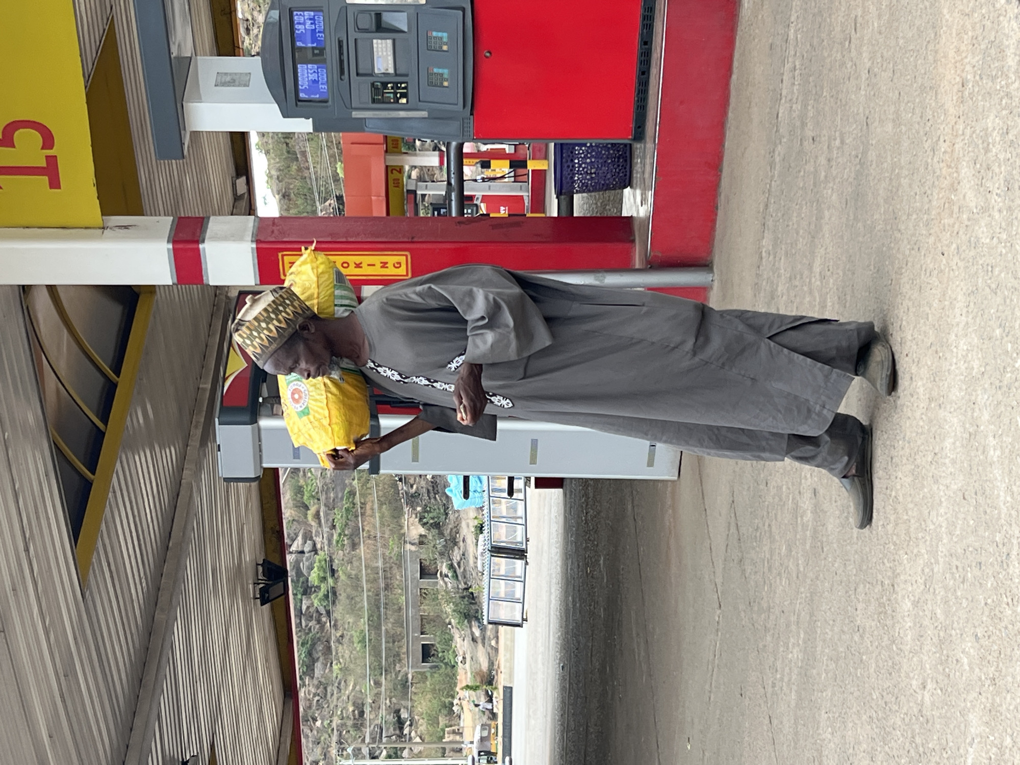 The image depicts a man wearing a traditional Nigerian cap and gray clothing standing next to a gas pump at a petrol station. The pump has a red canopy and a digital display showing fuel information. The man is holding a yellow bag in his left hand. The environment is urban or suburban with a distant view of rocky terrain. The location is Gura Top, Nigeria.