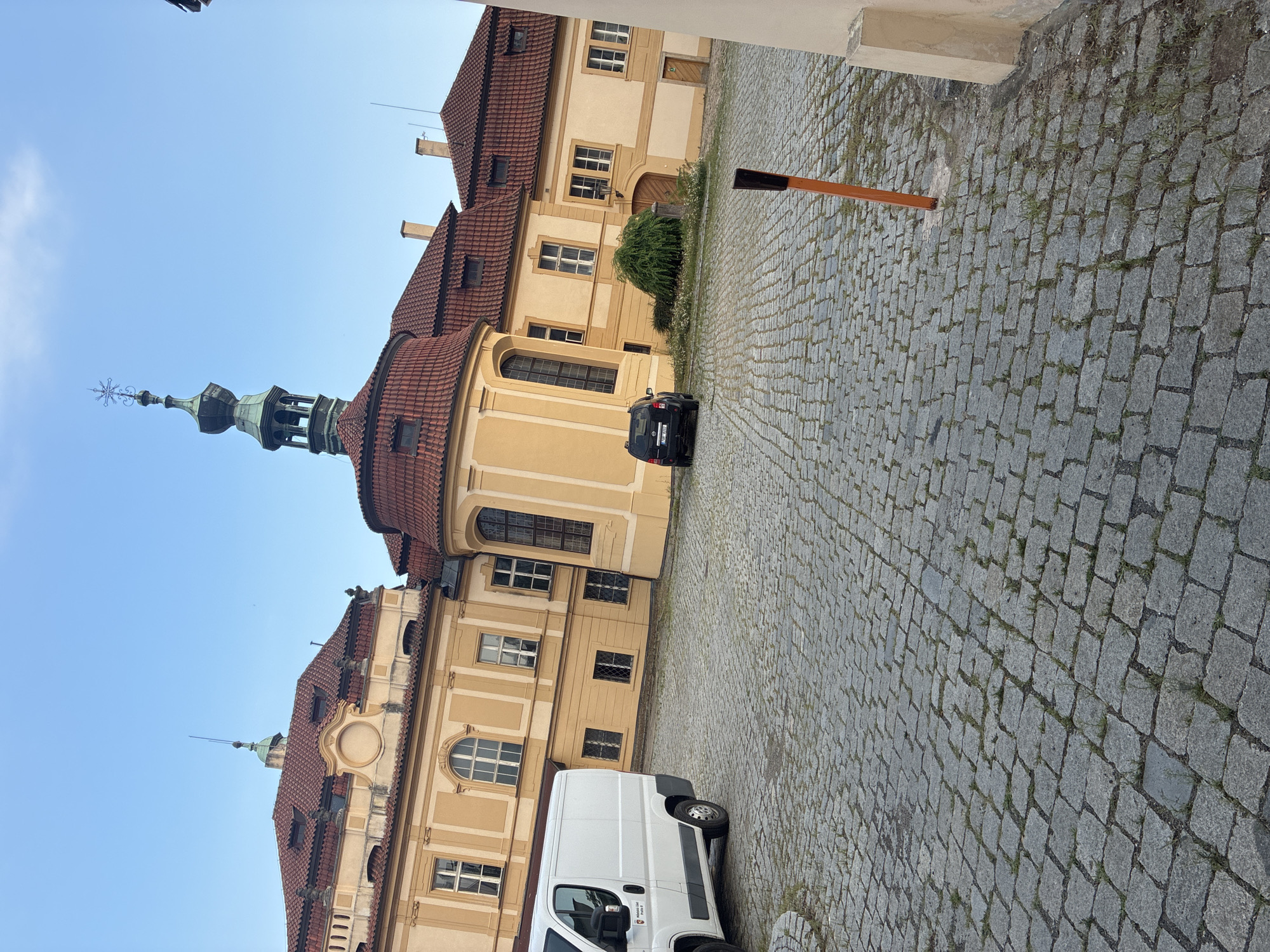 The image shows an exterior view of a building with a pale yellow facade and a red tile roof, characterized by its Baroque architectural style. The building features multiple windows, dormer windows on the roof, and a distinctive green tower topped with a cross-like finial. In the foreground, a cobblestone courtyard is visible, with a white van parked on the left and a black SUV parked on the right. The sky is clear and blue. This building is Strahov Monastery, located in Prague, Czechia, specifically in the Hradčany district. The scene is static, depicting a typical day in the courtyard of the monastery.