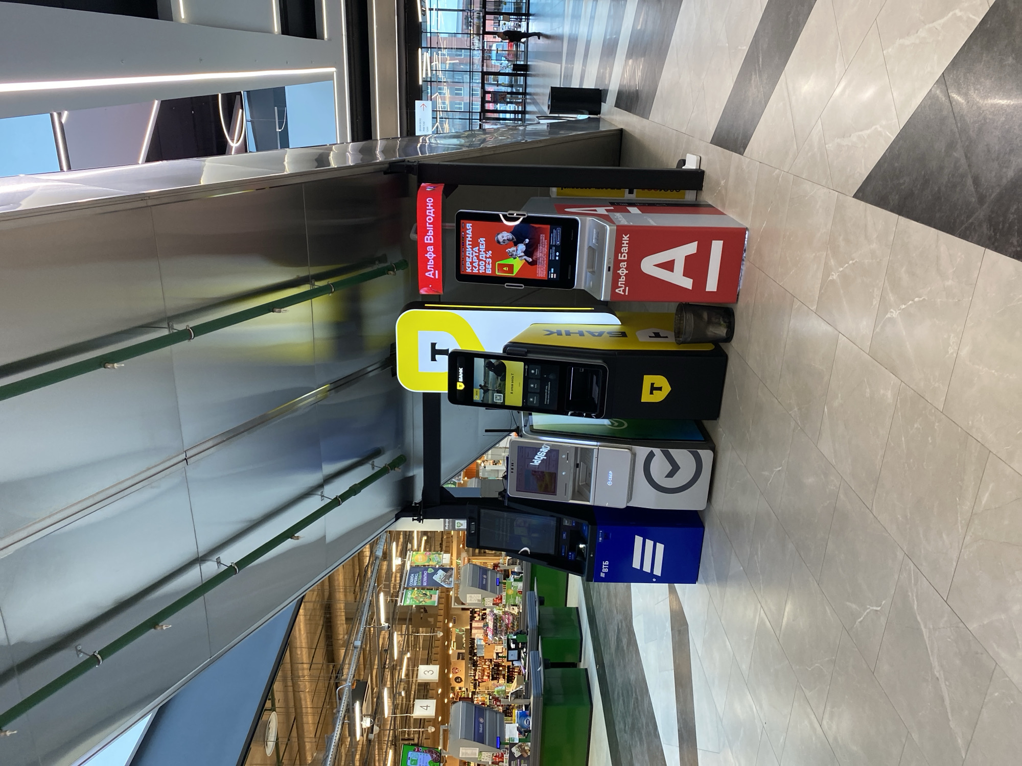 Here's a description of the image:
The image shows a row of automated teller machines (ATMs) or payment kiosks from various banks in an indoor environment, likely a shopping mall or similar commercial space in Murino, Russia.
The brands of the banks visible are Alfa Bank, Tinkoff Bank, VTB Bank, and Sberbank. The ATMs are lined up against a wall with a modern, angled architectural design. The flooring appears to be tiled with a light and dark gray pattern. In the background, a view into a shopping area with aisles and retail displays can be seen, suggesting the location is inside a commercial complex. The ATMs offer services related to banking, such as cash withdrawals or deposits, as well as various promotional services, such as credit card offers.