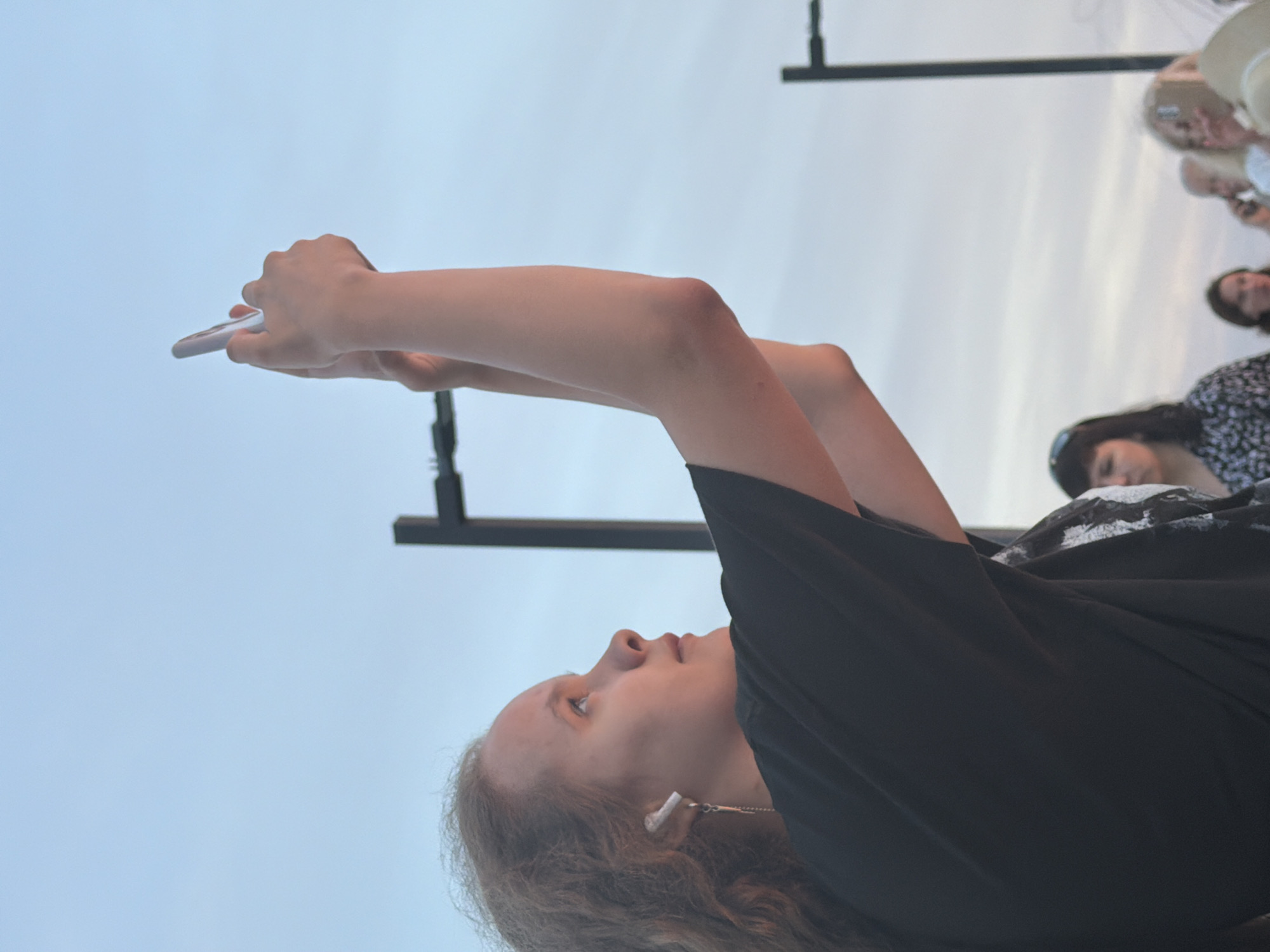The media file captures a close-up of a young person lying down and looking at their phone. The subject is positioned such that their arm is extended upwards, holding a white smartphone, while their head is tilted back, gazing at the screen. They appear to be wearing a black t-shirt and earbuds. The background is a light blue sky, suggesting it is daytime. In the upper right portion of the image, several other people are partially visible, some of whom are wearing hats. The overall scene seems casual and relaxed, possibly an outdoor gathering. There are no visible signs or text within the frame. The lighting and open sky suggest fair weather and daytime, likely late afternoon or early evening due to the soft light. The context provided places the scene in Sochi, Russia.