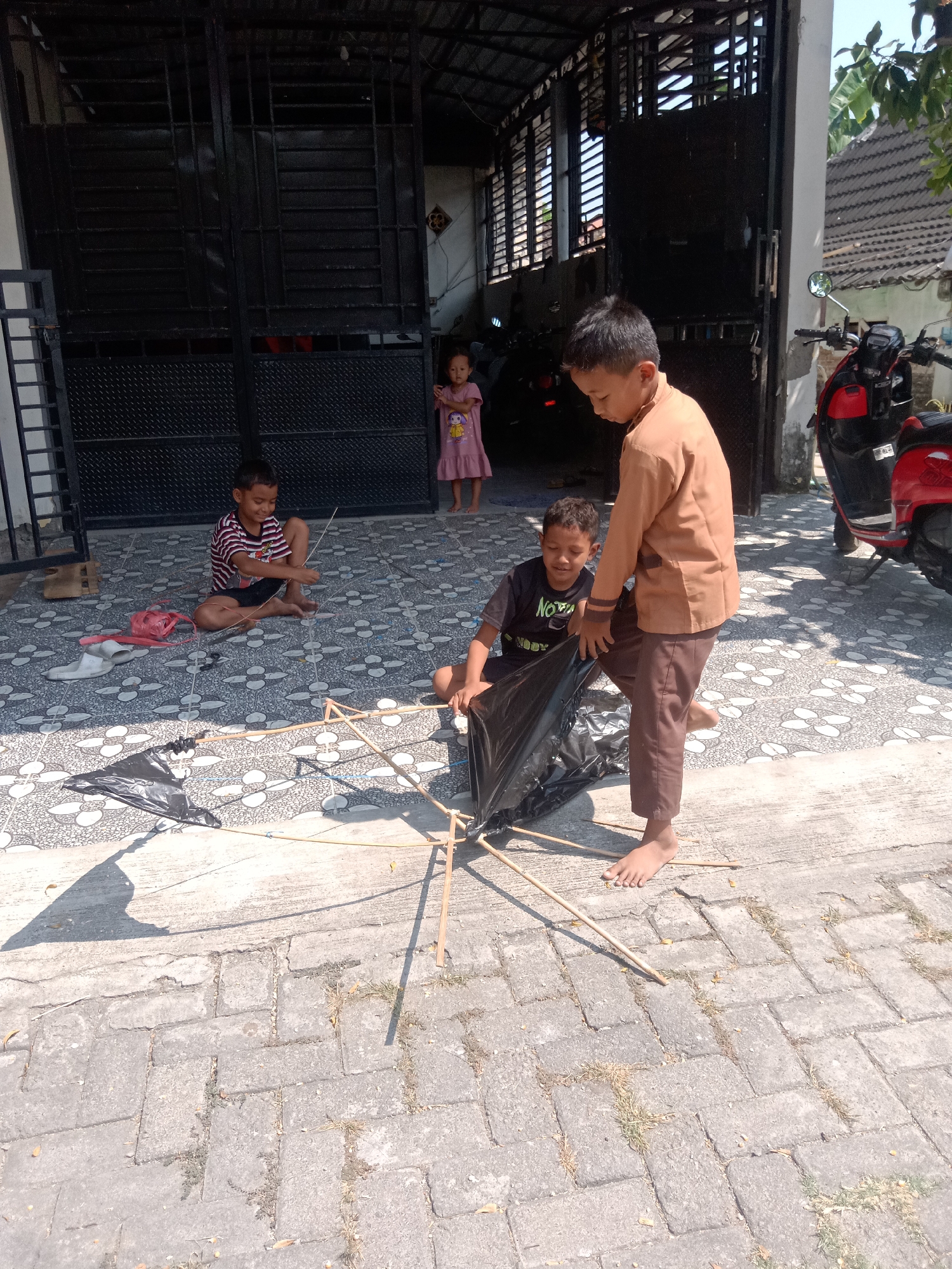 In the city of Stiaji, Indonesia, on a paved outdoor area, a group of children are engaged in assembling a kite. Two boys are actively working on the kite's frame, which is constructed from bamboo sticks and covered with black plastic. One boy, wearing a brown uniform shirt and pants, stands and appears to be guiding the process. The other boy, seated and wearing a black t-shirt, is also involved in holding and arranging the kite's materials. In the background, a third child, a young girl in a pink dress, stands near a dark metal gate, observing the activity. To the right, a red motorcycle is parked. The ground is a tiled patio, and sunlight casts shadows across the scene.