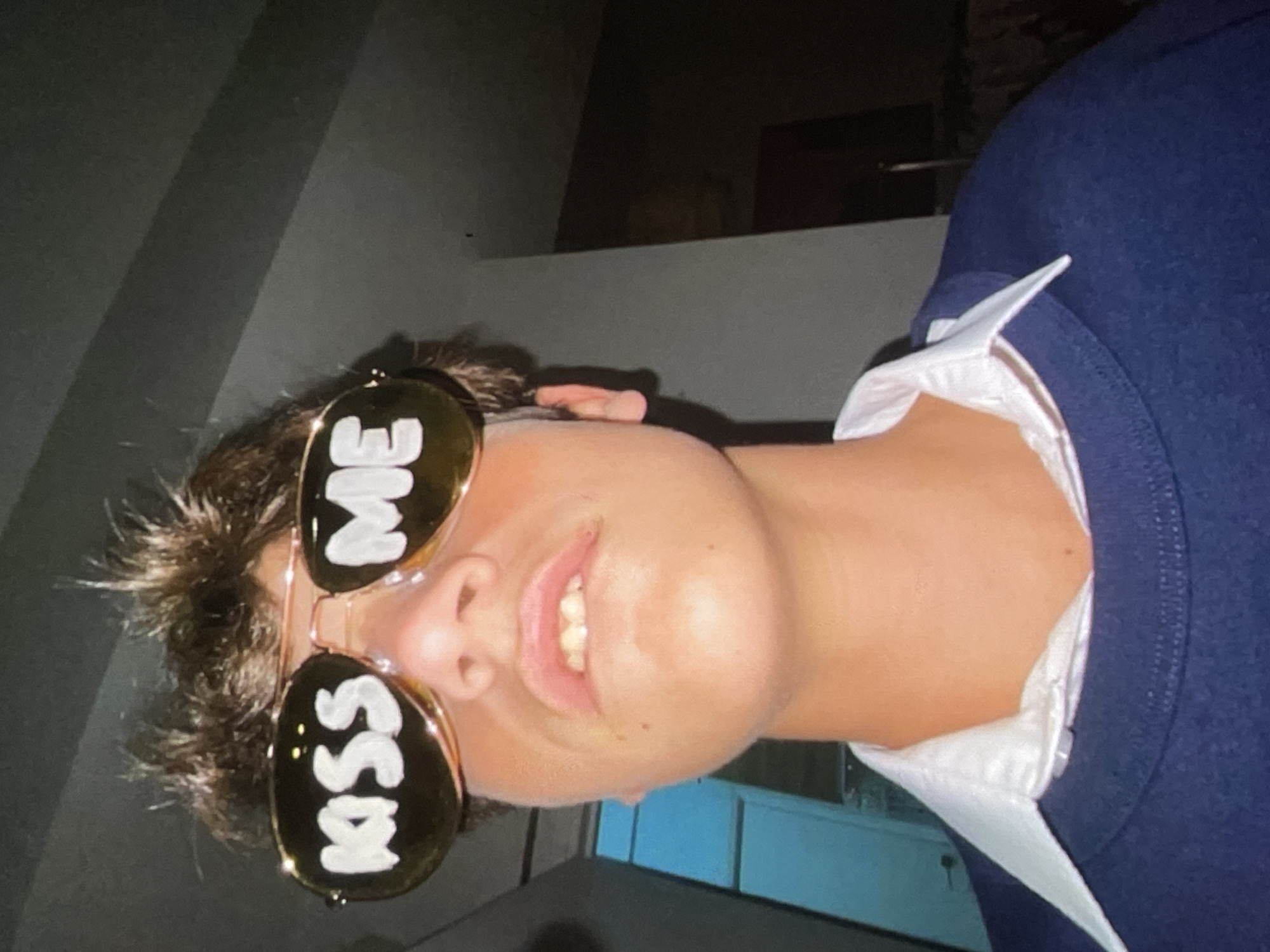 The image is a close-up, low-angle shot of a young person wearing aviator sunglasses with "KISS ME" written in white on the lenses. The person has brown, spiky hair and is wearing a navy blue sweatshirt over a white collared shirt. The background is dark and out of focus, suggesting an indoor setting. No specific location in Harmanli, Bulgaria, can be identified from the visual information provided.