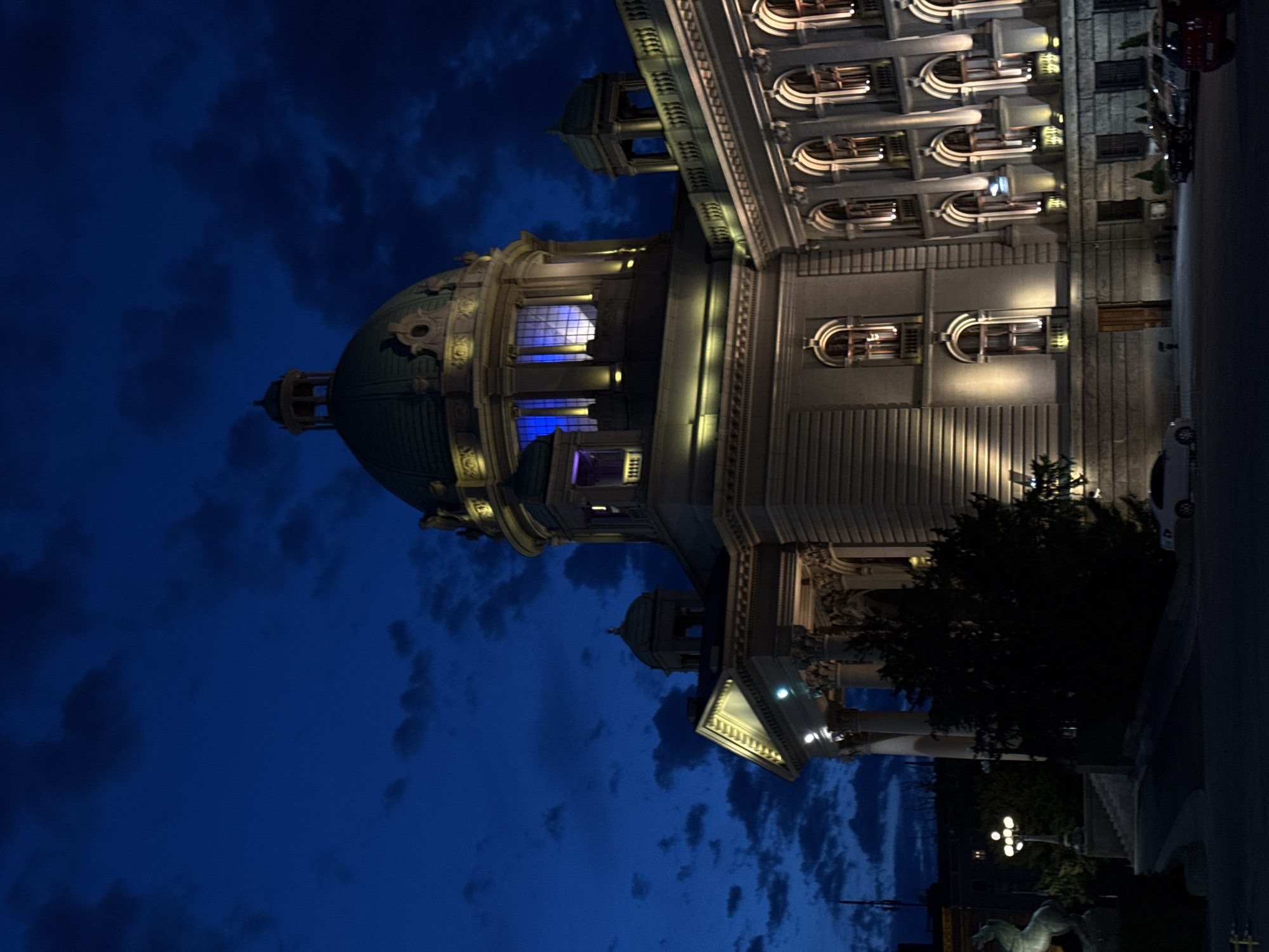 The image depicts the National Assembly of Serbia building in Belgrade, Serbia, at night. The building's architecture is highlighted with artificial lighting, emphasizing its dome and neoclassical design. The background features a dark blue sky with cloud formations. A street borders the building, showing parked vehicles and streetlights. Statues and landscaping are visible in the foreground. There are no people visible in the frame.