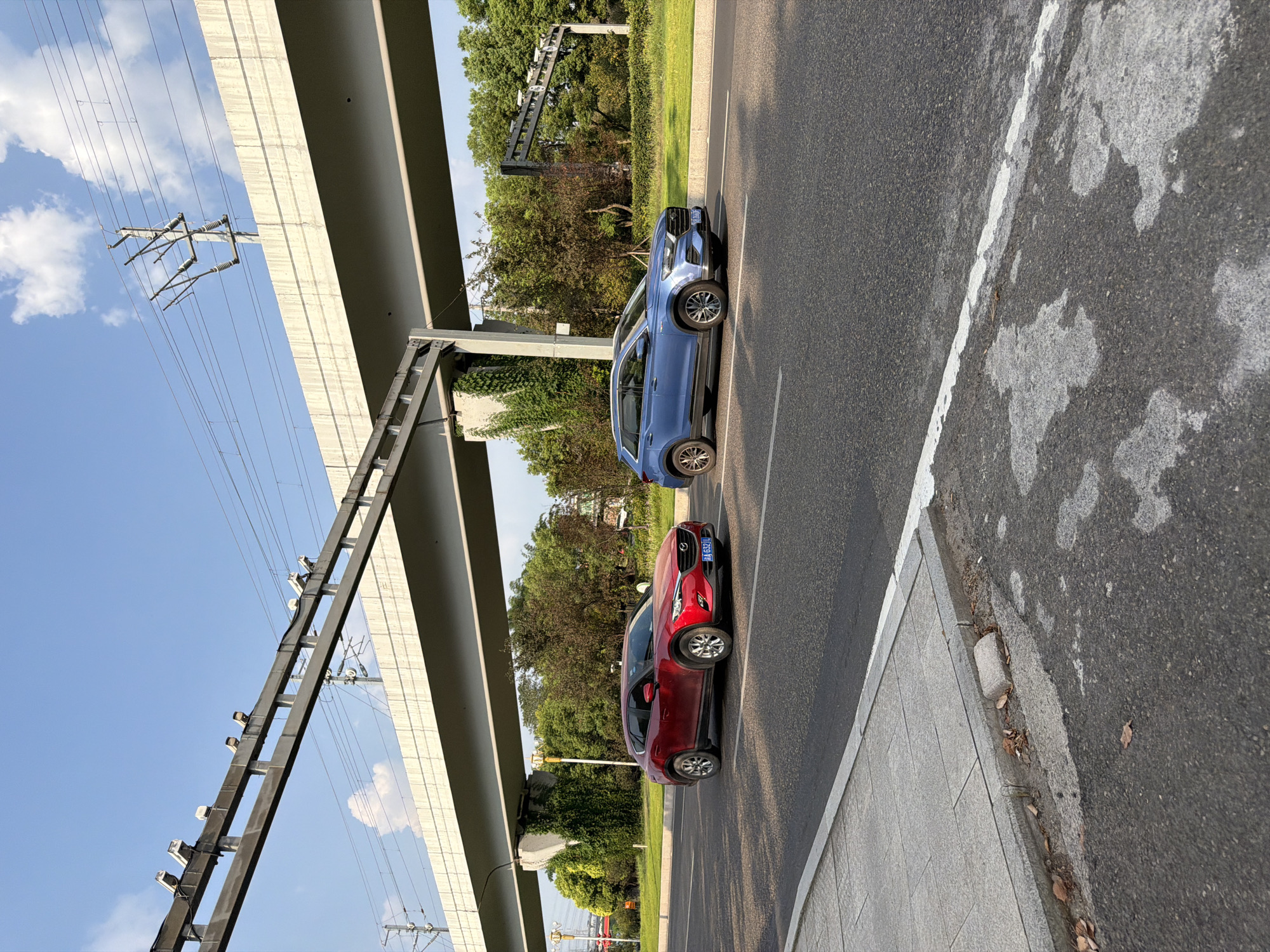 The image depicts a two-lane asphalt road in Jiangjin, China, under a large concrete overpass that supports overhead power lines and what appear to be traffic cameras. On the right lane of the road, a blue SUV is driving away from the camera. In the left lane, a red SUV is also driving away from the camera. The red SUV has a license plate that reads "JAA 632JL". A paved sidewalk with a concrete curb borders the left side of the road. The background features lush green trees and foliage, and the sky is clear with scattered white clouds. The lighting suggests it is daytime.