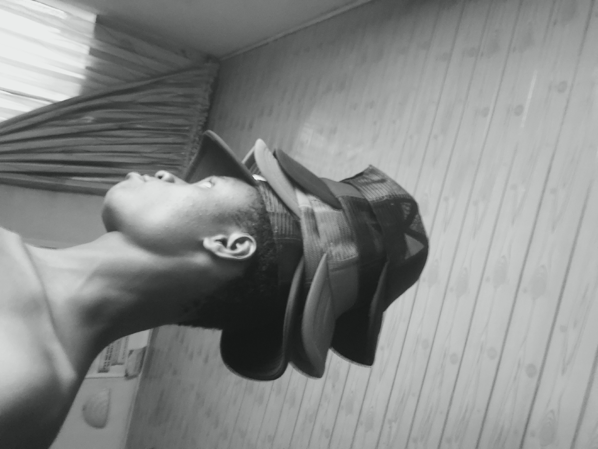 A black and white, low-angle, medium shot captures a shirtless individual with multiple baseball caps stacked on their head. The person is facing upwards, their neck exposed. The background includes a window with blinds and a wall with a wood-paneled design. An item is partially visible on the wall, perhaps a framed poster, slightly out of focus. The location is Narayi, Nigeria.