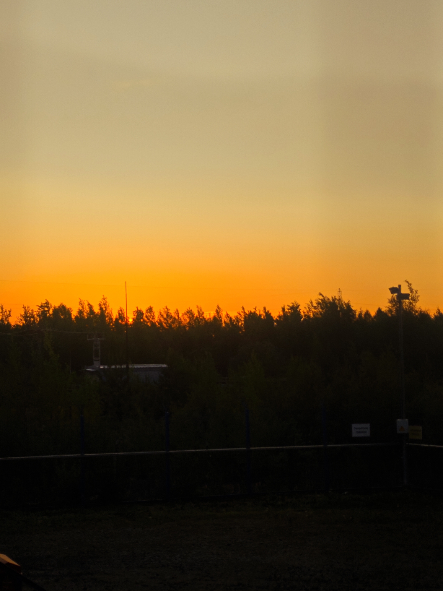A wide shot captures a sunset scene with a vibrant orange and yellow gradient filling the sky. A dark silhouette of a treeline stretches across the middle ground, with a small, indistinct building visible within it. A pole with a light fixture extends upwards from the treeline on the right side of the frame. A dark fence runs horizontally across the lower portion of the image, obscuring the ground beyond.