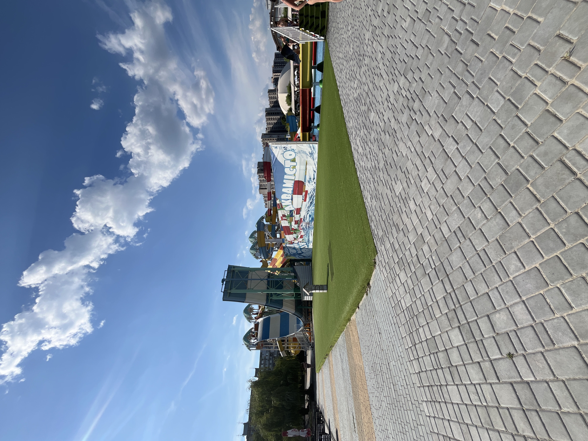 The image depicts a bright, sunny day at a water park in Dnipro, Ukraine. In the foreground, a meticulously paved walkway made of grey interlocking pavers leads towards a vibrant green area of artificial turf. Beyond the turf, a large water slide complex dominates the scene. One prominent slide is white with blue stripes, and another is a mix of red, yellow, and blue. A large banner displaying the text "АКВАMICTO" in white letters on a red and white background is visible. In the background, several modern apartment buildings are situated. A large willow tree stands to the left of the water slides. People are visible near the slides and on the walkways, suggesting the park is open and active. The sky is a clear blue with scattered white clouds.