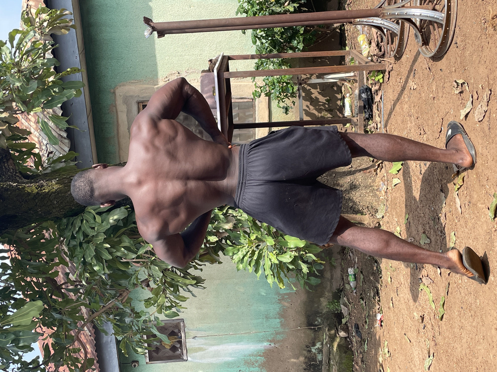 A full-body, rear-facing shot captures a muscular Black man standing outdoors in Akwanga, Nigeria. He is positioned in front of a green, weathered building with a corrugated metal roof and a small, barred window. A large tree trunk is to his left, with lush green foliage and some visible fruits. The man is wearing black athletic shorts and dark sandals. His hands are on his hips, and his back muscles are prominently displayed. The ground is dirt, littered with dry leaves and some discarded plastic bottles. In the background, a rusty metal table stands to his right, and further to the right are parts of what appear to be metal structures, possibly scrap or equipment. The lighting suggests it is daytime, with shadows cast on the ground.
