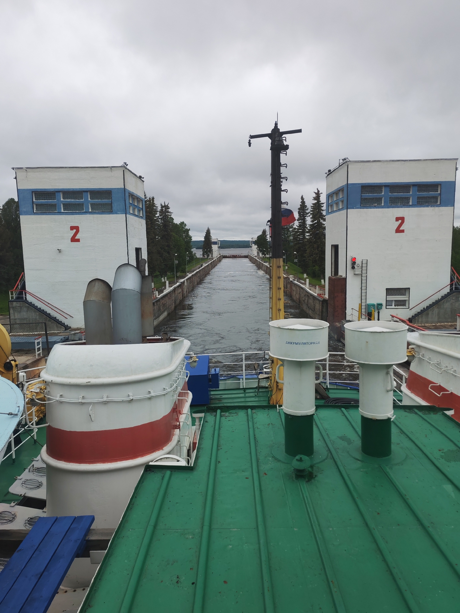 The image captures a scene at lock number two on the Volga–Don Canal, likely near the city of Белоусово, Russia. It is taken from the deck of a boat moving through the lock. The canal stretches out between two control towers marked with the number "2" in red. Each tower is a two-story, white building with a horizontal blue stripe beneath the roofline. The sky is overcast, and evergreen trees line the canal banks. The canal walls are constructed of concrete.
In the foreground, the green-painted metal roof of the boat dominates, featuring ventilation shafts. A portion of the boat's superstructure is visible, painted in white with a red stripe. A blue wooden bench rests on the edge of the roof. A mast is visible.