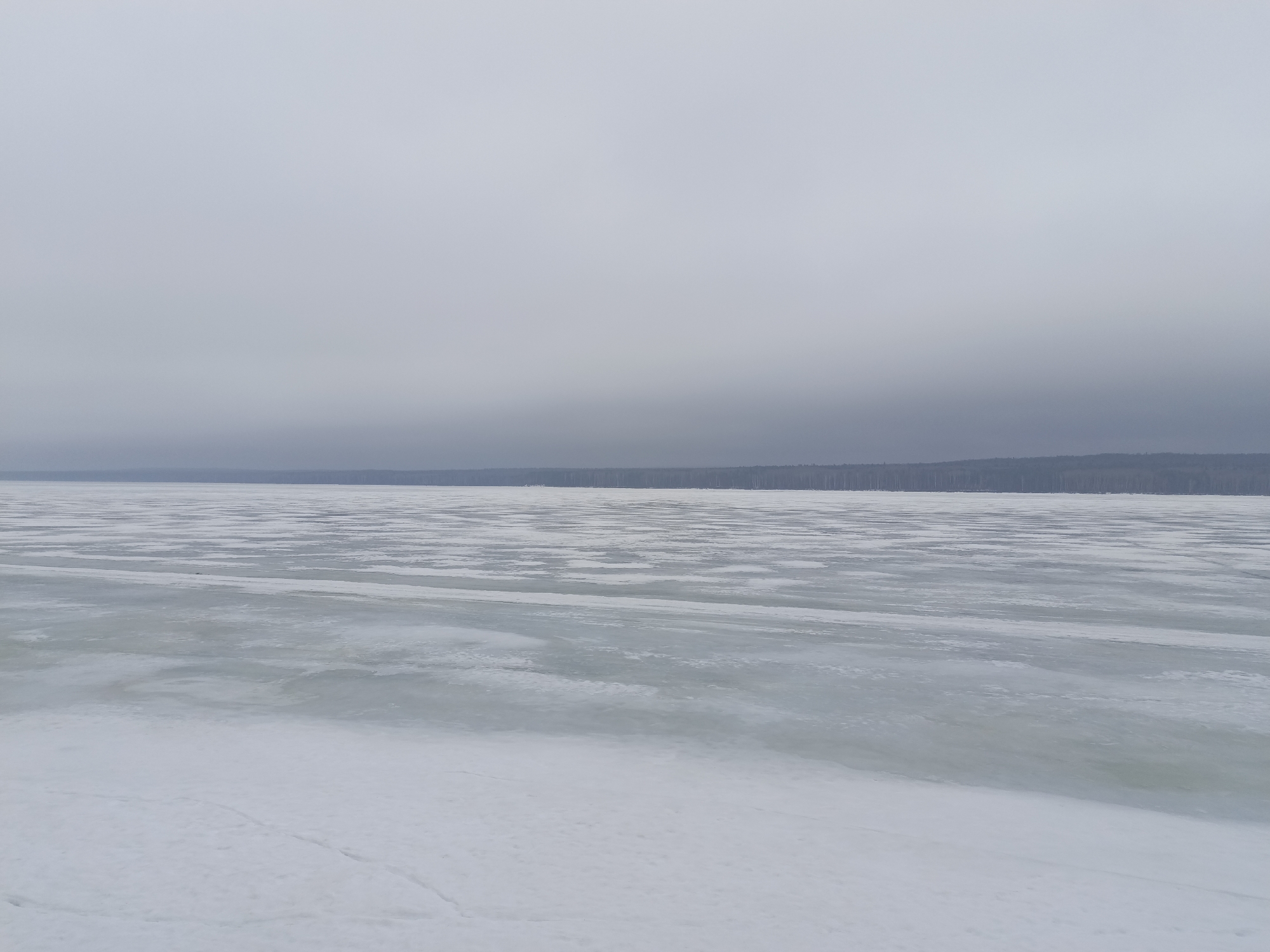 The image shows a frozen lake or large body of water under an overcast sky. A line of trees can be seen in the distance along the opposite shore. The ice on the lake has a patchy appearance, with areas of smooth ice interspersed with areas of rougher ice and snow. The location is likely near the city of Chaikovsky, Russia, given the prompt's specification. There are no people or other visible objects in the scene besides the natural elements.