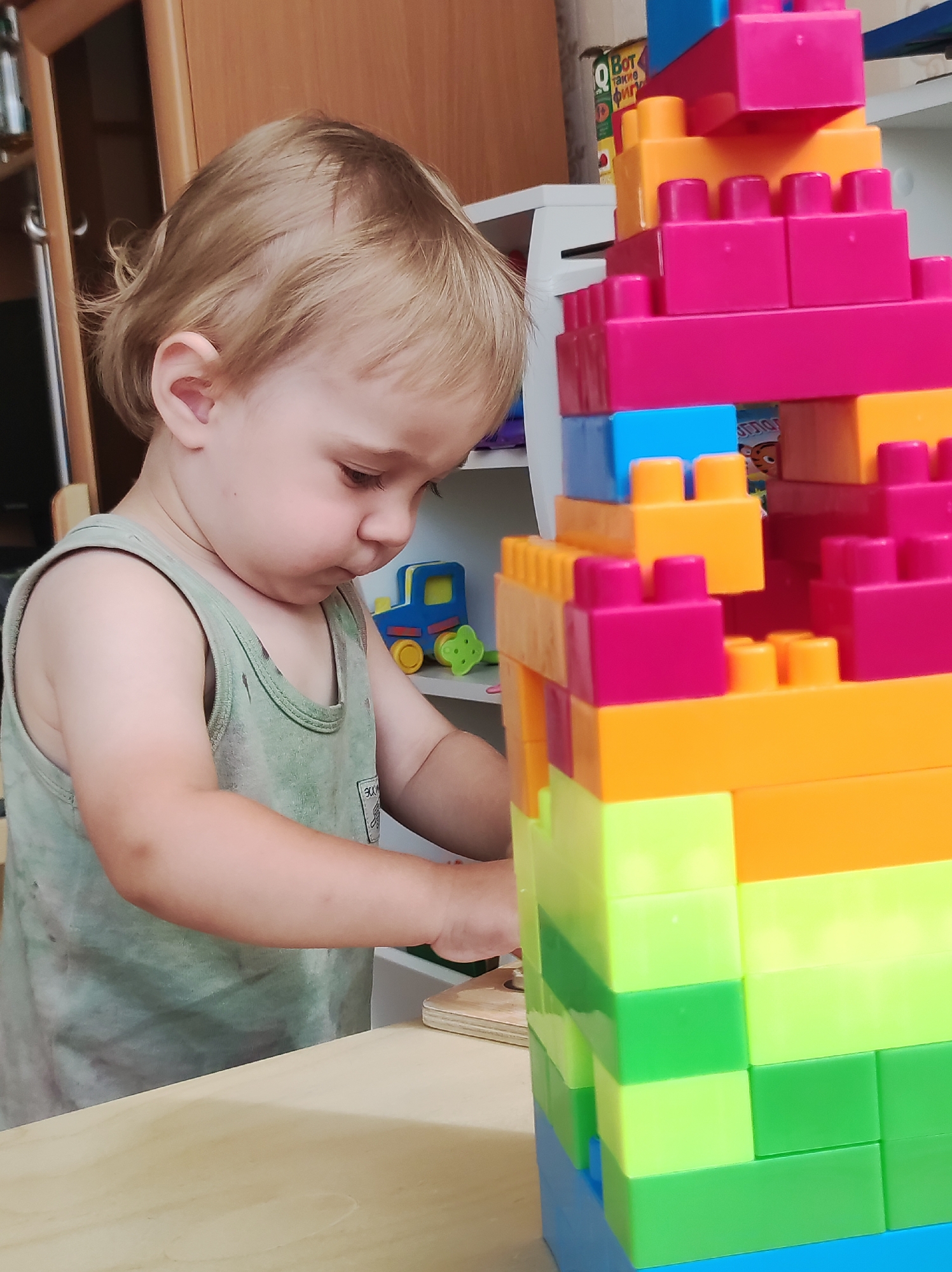 In a residential interior, likely in Chaikovsky, Russia, a young child with light brown hair and fair skin, wearing a green tank top, is engrossed in building a tall, multi-colored tower of interlocking plastic blocks. The tower, predominantly composed of pink, orange, light green, and blue blocks, stands to the child's right. In the background, a white shelf is visible, with a blue toy truck with yellow wheels on it. The scene appears to be a domestic play area.