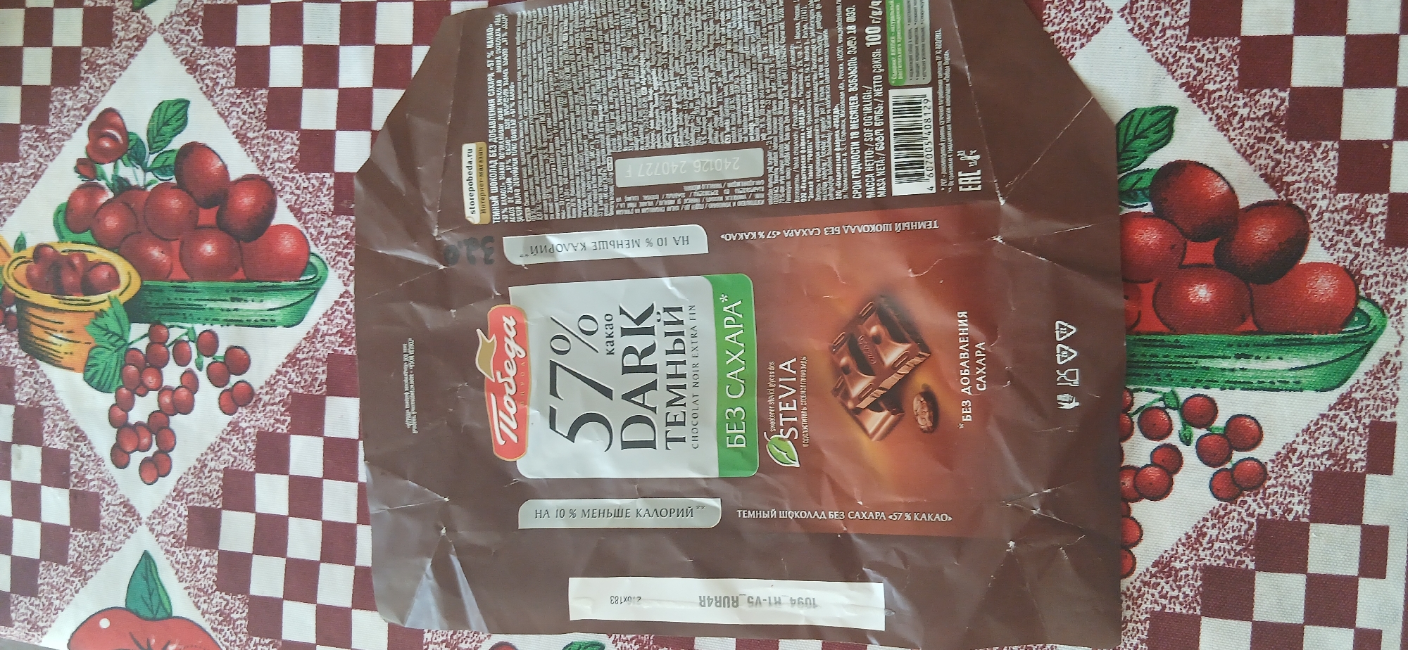 The image displays the crumpled wrapper of a "Pobeda" brand dark chocolate bar. The wrapper features a prominent "57% Cacao" designation, along with "Dark Temnyy" and "Chocolat Noir Extra Fin." It also highlights "Bez Sakhara" (without sugar) and mentions Stevia sweetener. The background of the image is a red and white checkered tablecloth with a fruit pattern, suggesting it might be on a table. There are no people or discernible elements that would indicate a specific location in Andijan, Uzbekistan. The packaging is the sole subject of the photograph, presented in a close-up view.