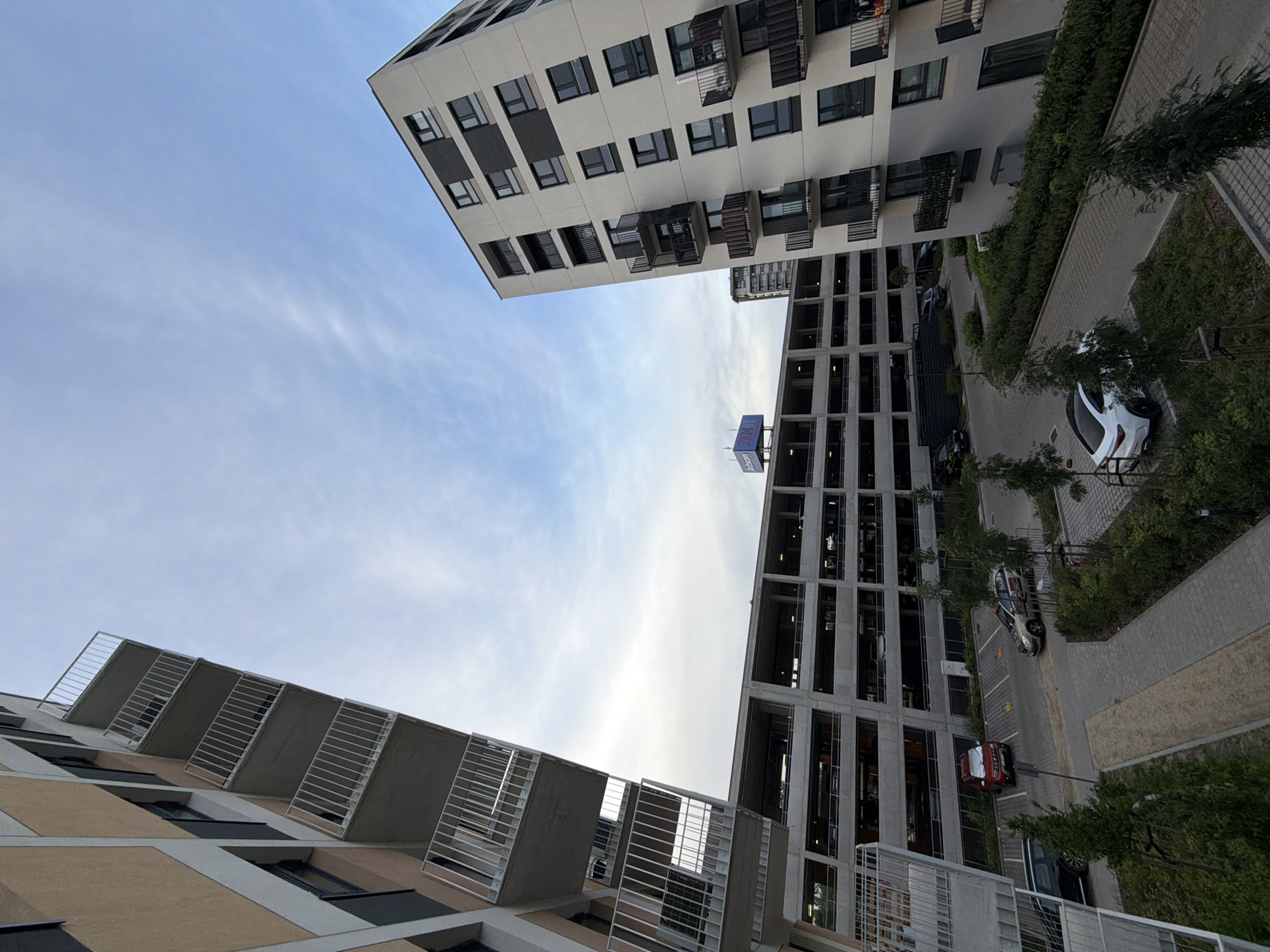 The image shows a view of modern apartment buildings and a multi-story parking garage in the city of Łódź, Poland. The buildings feature contemporary architectural designs with balconies and large windows. The parking garage is constructed from concrete and appears to be several levels high, with cars visible on some of the levels. Several cars are parked in an outdoor lot adjacent to the buildings, including a white sedan, a taxi, and a red SUV. The sky above is partly cloudy with a pale blue hue, suggesting it is either early morning or late afternoon. Greenery, including trees and shrubs, surrounds the parking area and the base of the buildings, indicating a planned urban landscape. A sign with the word "ZENITH" is visible on top of the parking structure.
