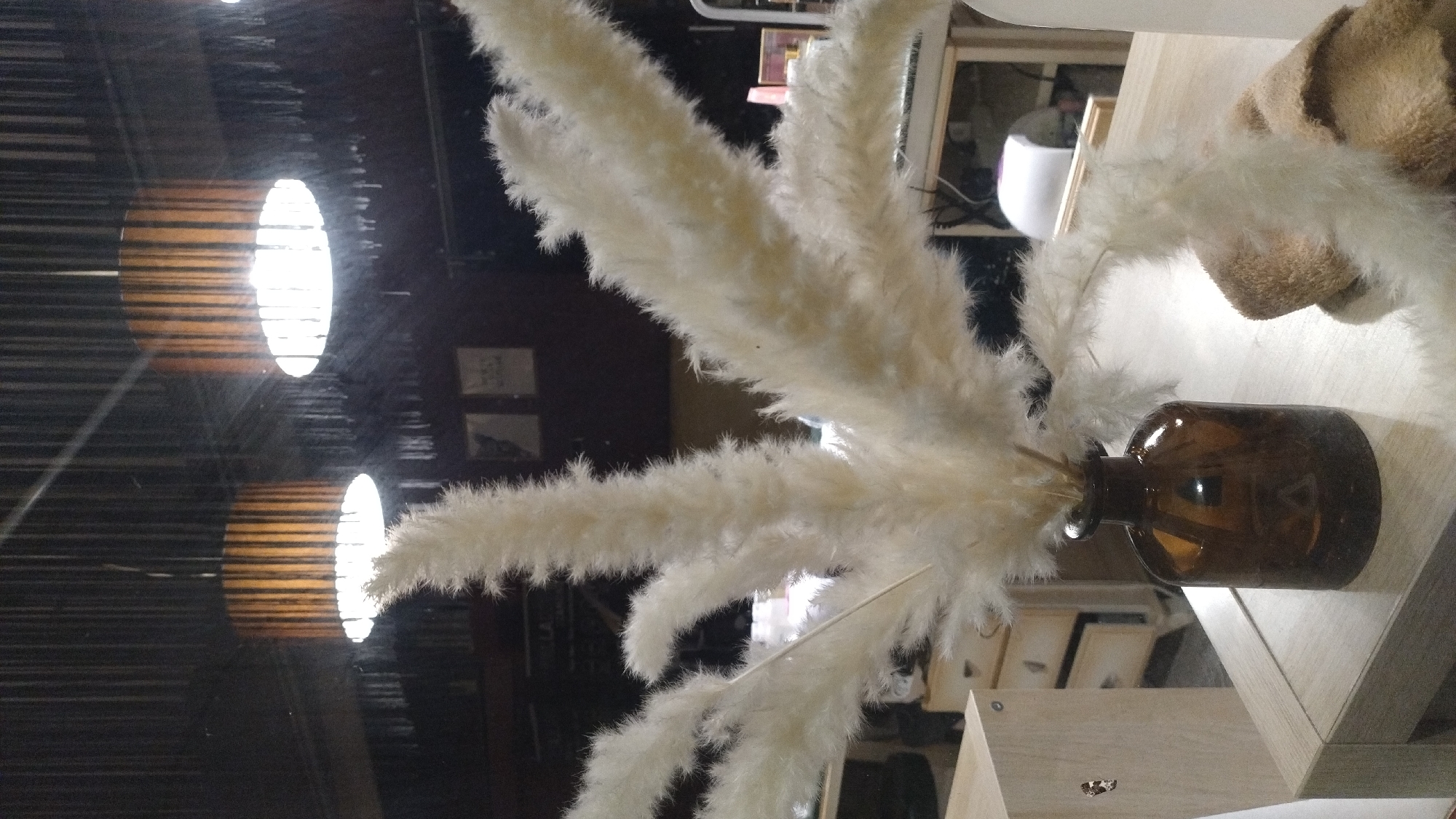 In Perea, Greece, an indoor scene depicts a cluster of dried, fluffy pampas grass stalks arranged in a dark amber glass vase. The vase rests on a light-colored wooden table. In the background, two pendant lights with slatted wooden shades hang from a dark ceiling. The environment appears to be a room with some furniture, including a light-colored cabinet with drawers to the left and what looks like a desk or workbench with equipment in the mid-ground to the right. The lighting is dim, with the pendant lights providing the primary illumination. There are no people visible in the image.