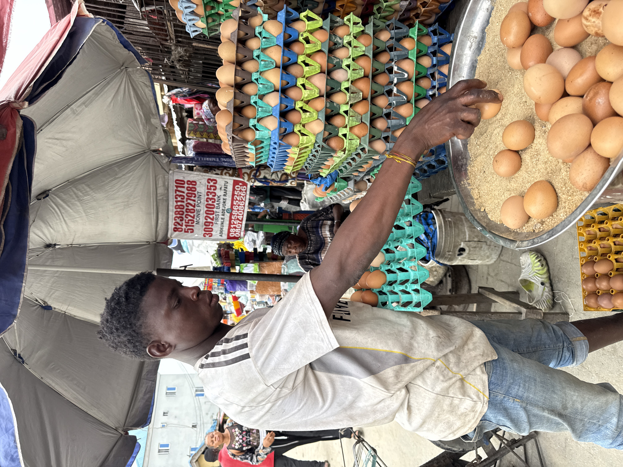 The scene shows a street vendor in Ado, Nigeria selling eggs. The vendor, a young black man, is picking up an egg from a metal basin filled with eggs and wood shavings. Stacks of egg cartons in various colors (blue, green, yellow) are visible behind him. An umbrella provides shade. A sign displays financial transaction information, including phone numbers, bank details, and a name, "ADEKUNLE ADEJOKE RAFATU" alongside "FIRST BANK." In the background, other people and various merchandise are visible, indicating a bustling marketplace.