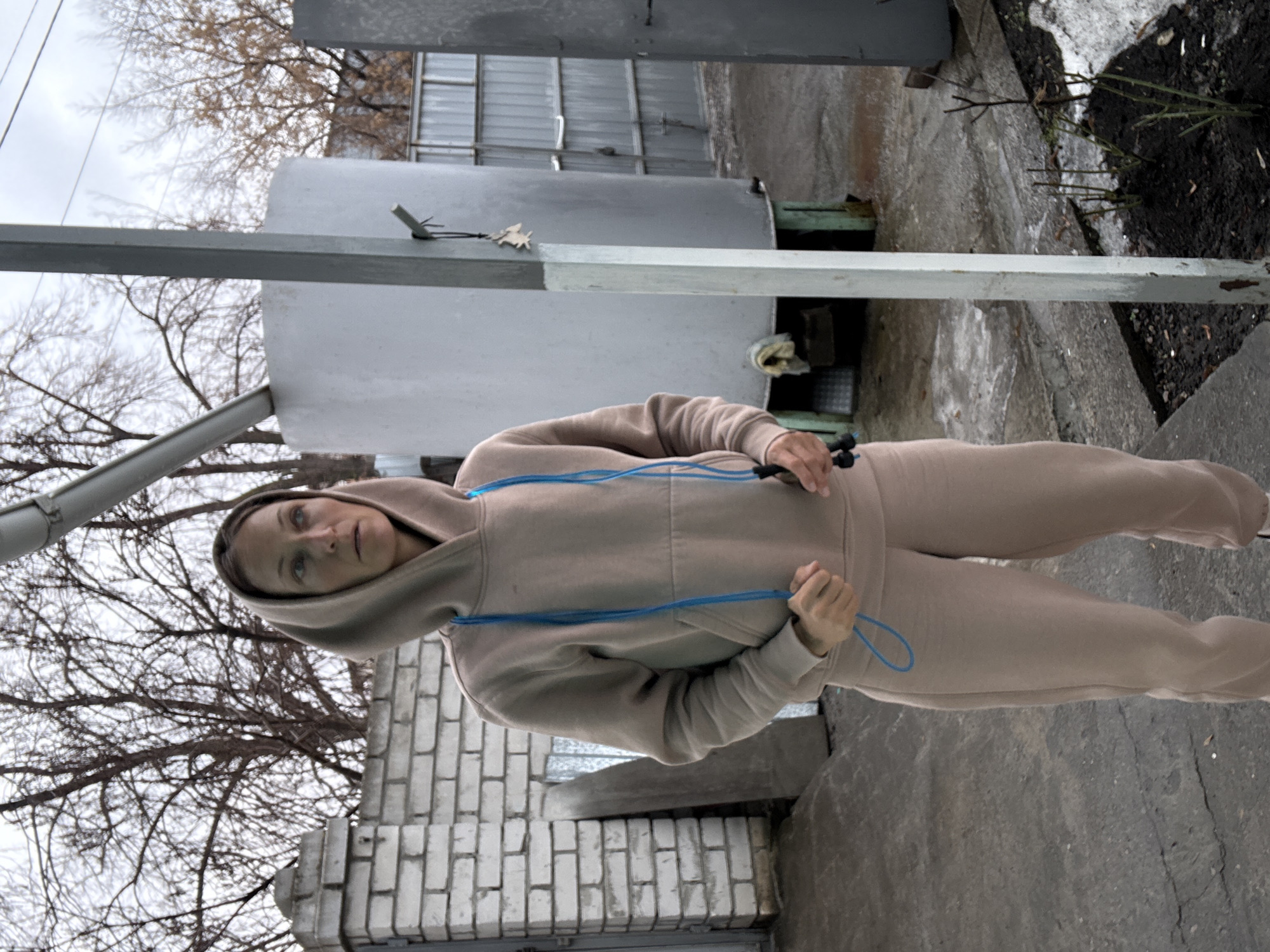 A woman stands in an outdoor location in Ulyanovsk, Russia. She wears a beige sweatsuit with a hooded top and is holding a blue jump rope. The background features a gray metal structure, bare trees, a brick wall, and concrete pavement. The sky appears overcast.