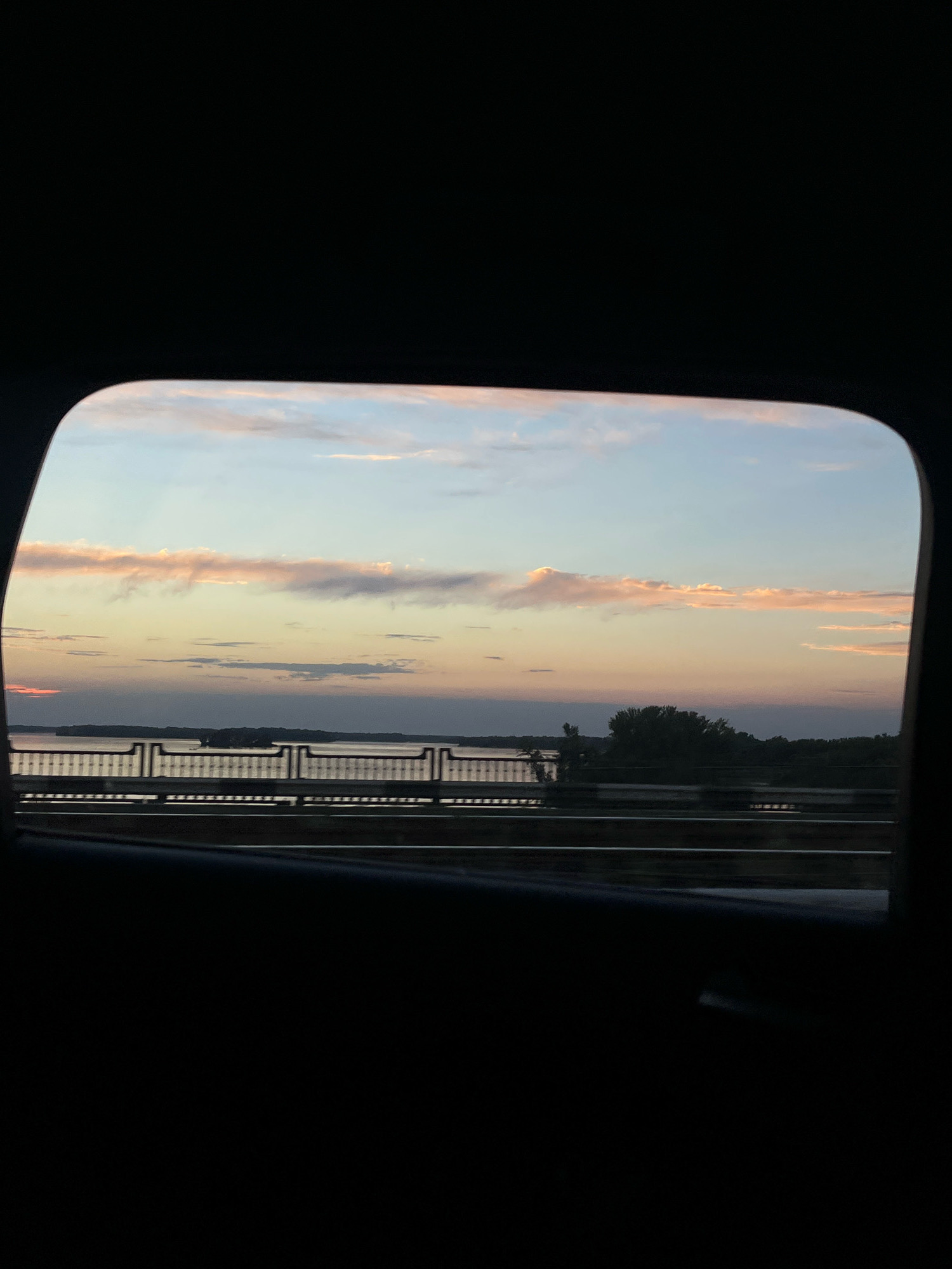 A view through a car window shows a tranquil sunset over a body of water. The sky is a gradient of soft blues, pinks, and oranges, with wispy clouds scattered across it. A distant shoreline with silhouetted trees can be seen. In the foreground, a metal railing separates the viewer from a road or bridge. The bottom portion of the image is dark, indicating the interior of the car.