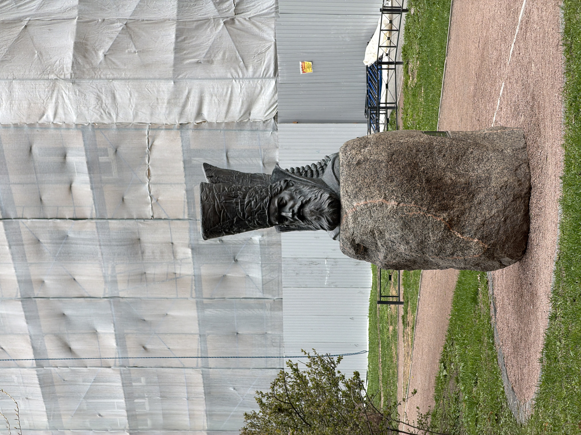 The image shows a sculpture in Saint Petersburg, Russia. The sculpture, depicting a man's head, is made of dark metal and sits atop a large, rough stone base. The sculpture and base are positioned on a narrow path between a grassy area and a pebble-covered walkway. Behind the sculpture, a building is covered in white protective material, suggesting ongoing construction or renovation. To the right of the sculpture, metal supports and a partially visible structure stand along the path. In the foreground, the branches of a small tree extend into the frame.