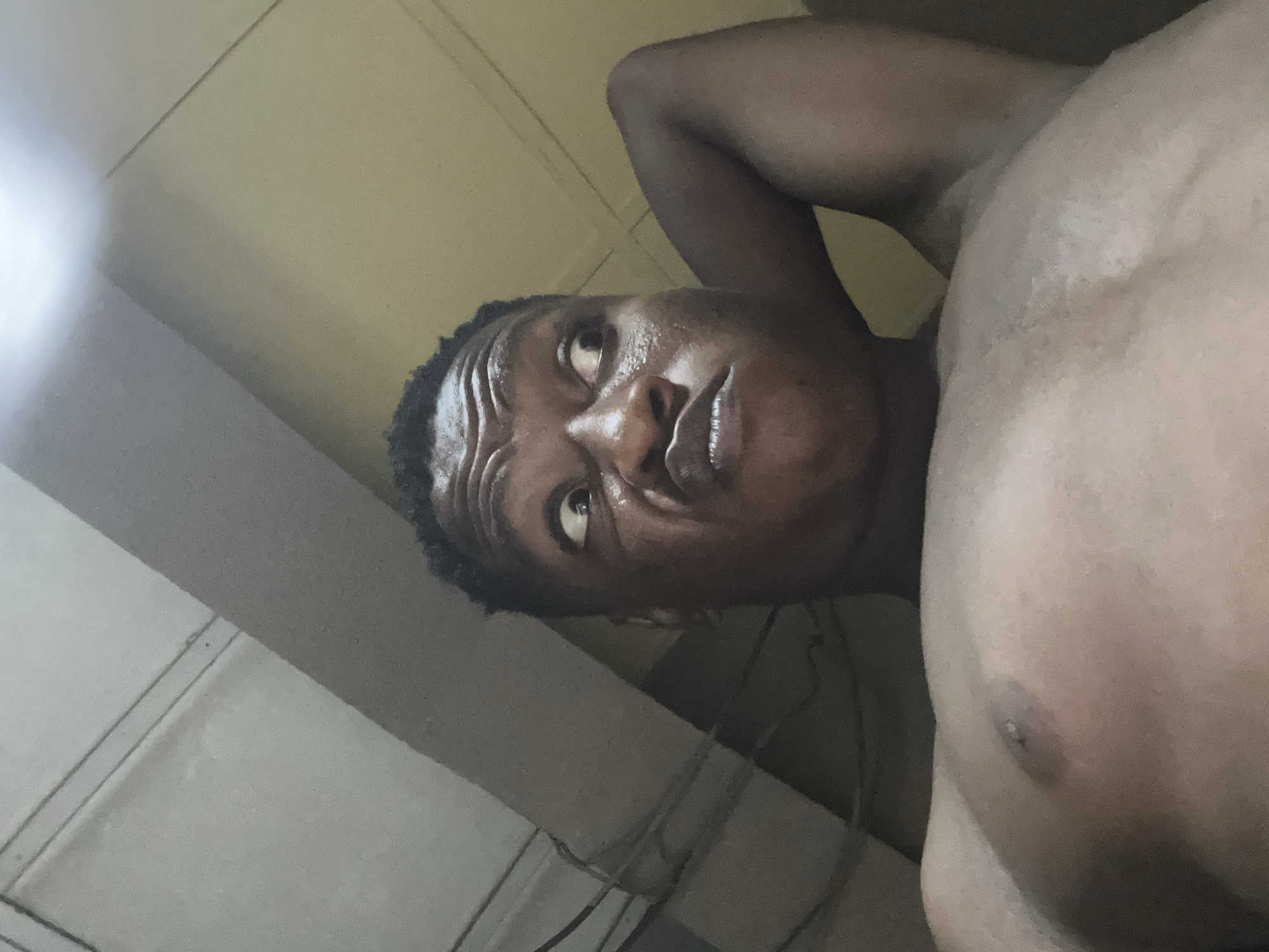 The image displays a close-up, low-angle shot of an adult male of African descent, photographed indoors in what is contextually identified as Ikorodu, Nigeria. The individual is shirtless, occupying the central and lower portions of the frame. His head is tilted slightly back, and his gaze is directed upwards and to the right with wide, possibly surprised or bewildered eyes. His forehead is deeply furrowed with horizontal lines, and his mouth is pursed and scrunched, indicating a strong facial expression. His left arm is bent at the elbow, with the hand positioned behind his head, and the elbow pointing towards the upper right of the frame. The subject's bare chest, including visible nipples, is present in the lower part of the image.
The background depicts an interior room with muted colors. The upper left portion shows a light grey ceiling or wall section with two thin, dark parallel lines, possibly electrical conduits or pipes. A darker grey, diagonal structural element or beam runs from the upper left towards the center-right. The upper right portion of the background features a lighter beige-colored wall or ceiling, illuminated by a bright, diffused light source originating from the upper right corner. The overall lighting is dim, characteristic of an artificially lit indoor environment. The presence of visible conduits and plain wall surfaces suggests a functional or residential setting.