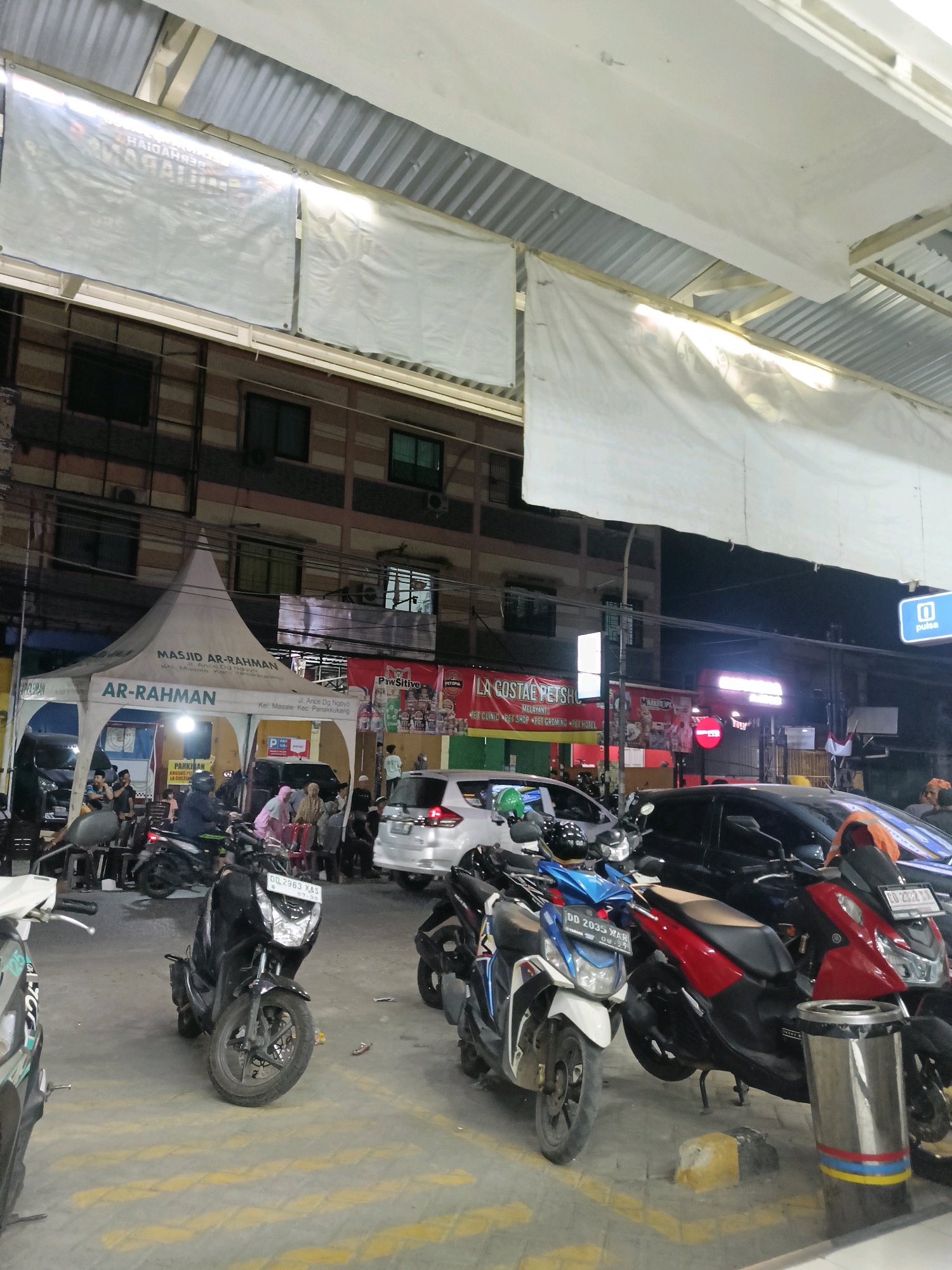 This is a nighttime, eye-level, medium shot of an outdoor street scene in Makassar, Indonesia, likely a marketplace or street vendor area. The foreground features several parked motorcycles, including a black scooter with the license plate "DL 2963 XAS" and a white and blue scooter with the license plate "DD 2035 XAR." To the right of these scooters is a red and black motorcycle and a trash can. In the midground, a white tent with "MASJID AR-RAHMAN" and "AR-RAHMAN" printed on it is set up, with people gathered underneath it. Behind the tent, a white car is parked. Further back, there are buildings with multiple windows and balconies. Signs are visible on the buildings, including one that reads "LA COUSTAE PETSHOP" and another with "PULSA." Several people are visible throughout the scene, some standing and some seated. The overall environment appears to be a busy commercial area at night, with artificial lighting illuminating the scene. The ground is paved, with yellow lines marking parking spaces. The background shows a dark sky, indicating it is nighttime.